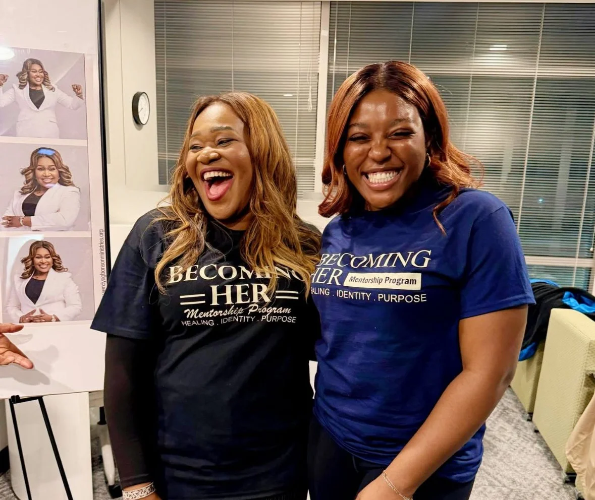 Two women smiling and posing together at a mentorship program event. They are wearing matching T-shirts with the words 'BEcoming HER Mentorship Program' and the phrase 'Healing, Identity, Purpose.' Behind them is a poster with photos of the same woman in different poses.