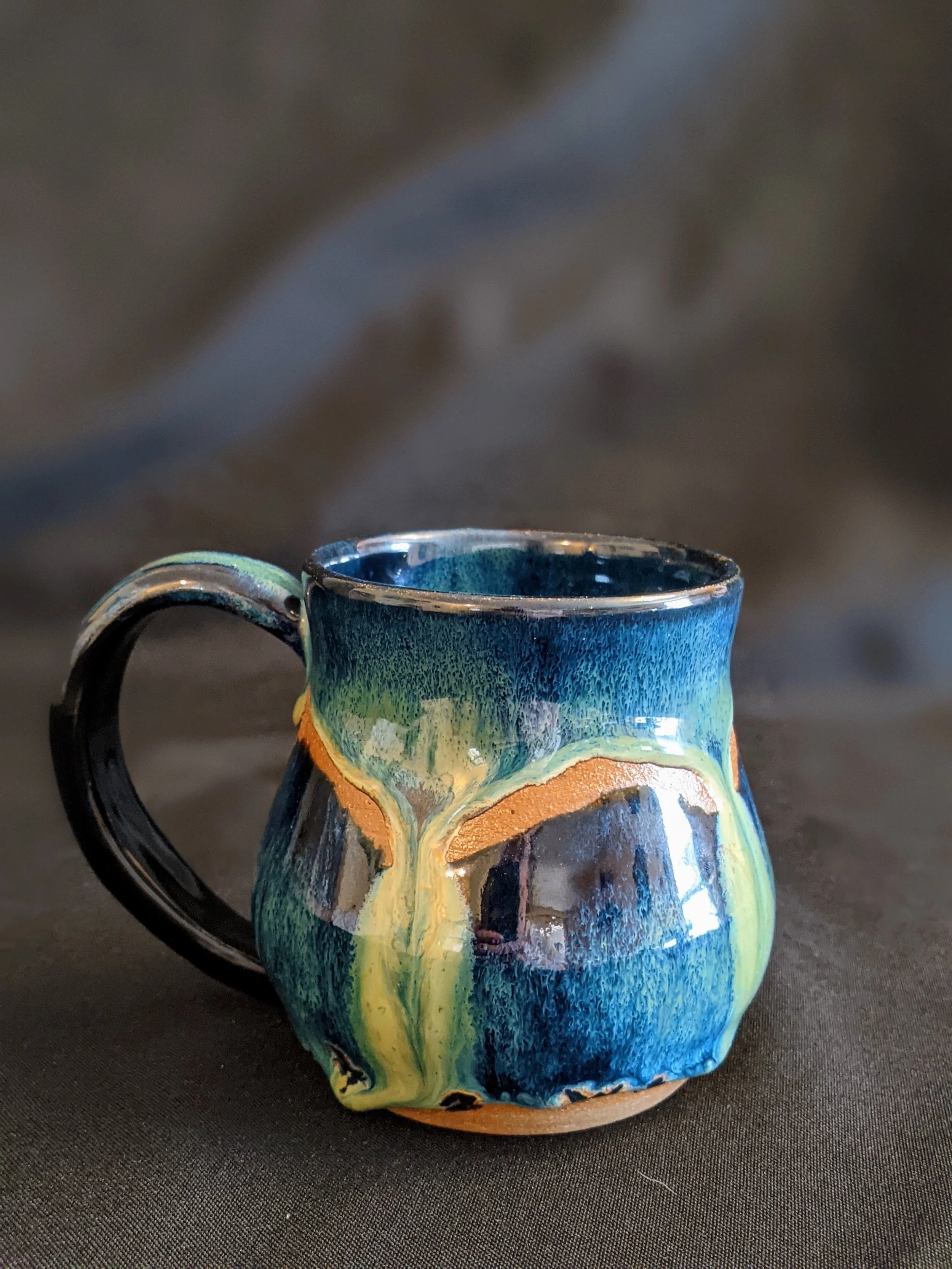 A colorful ceramic mug with abstract paint designs in blue, green, orange, and black, placed on a black surface.