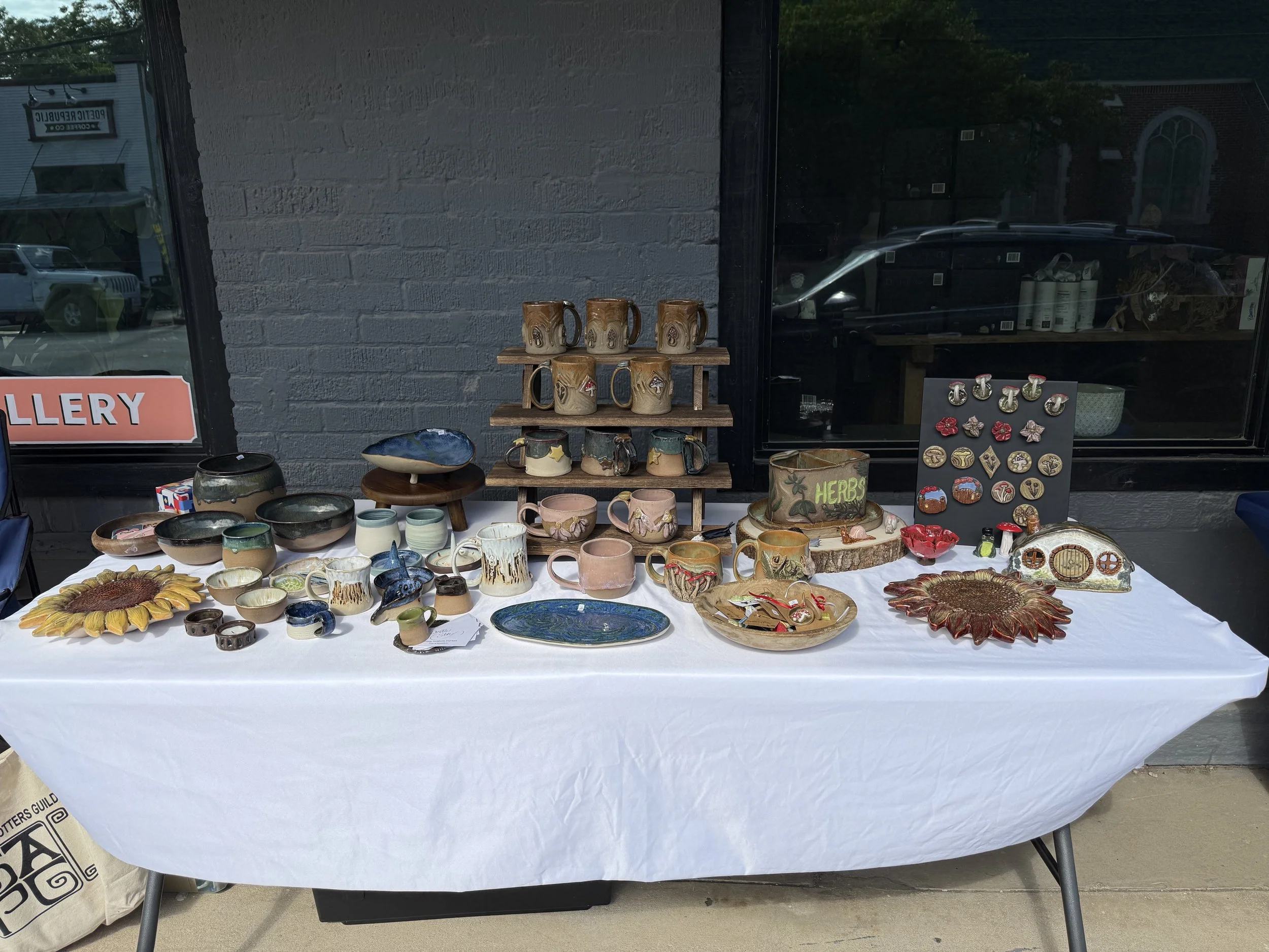 A table of pottery and ceramic items, including mugs, bowls, and plates, displayed outside a storefront with a gray brick wall and glass window behind it.