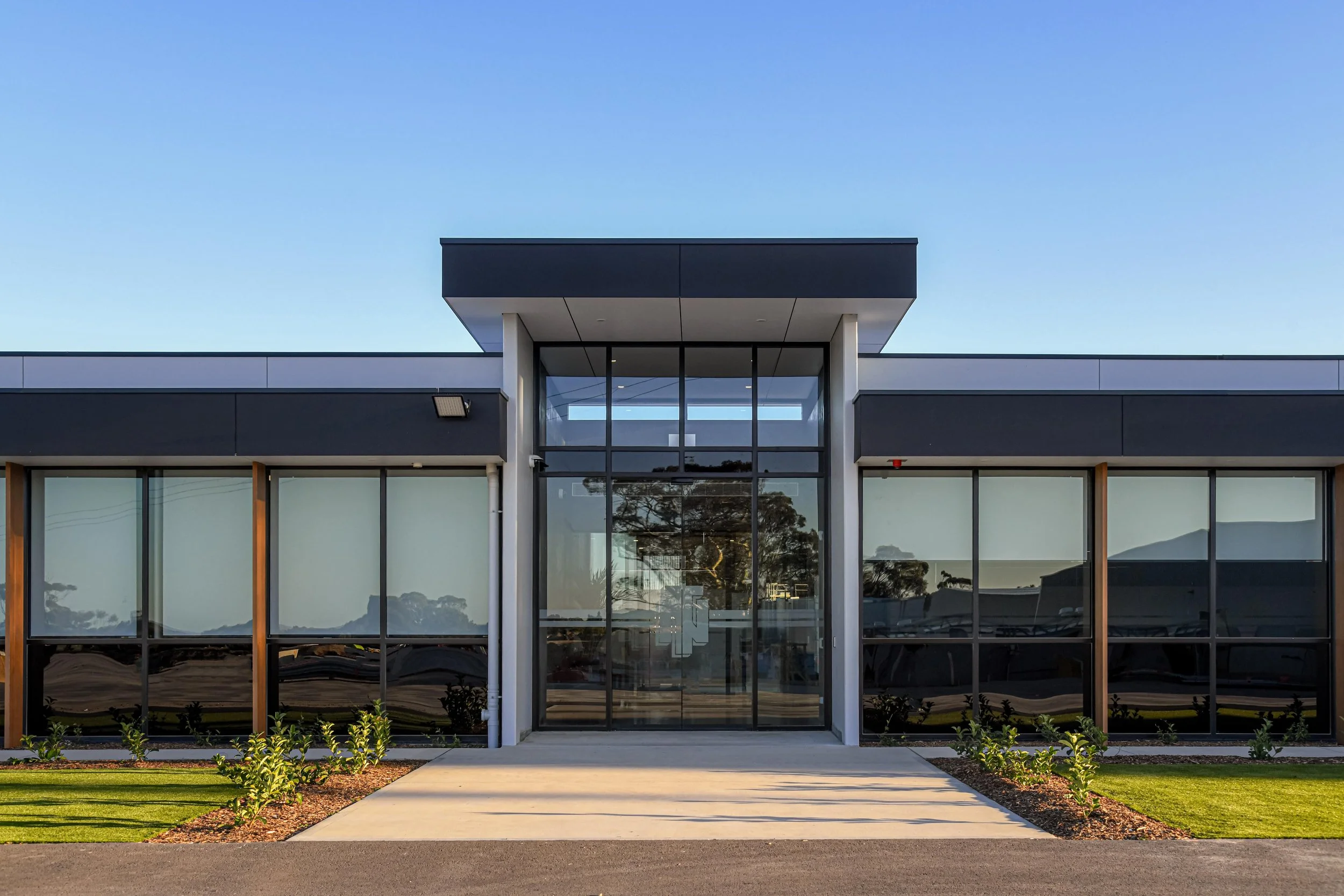 Front view of Nuriootpa Traders new office, with large glass windows, surrounded by a concrete walkway, green landscaping, and a clear blue sky.