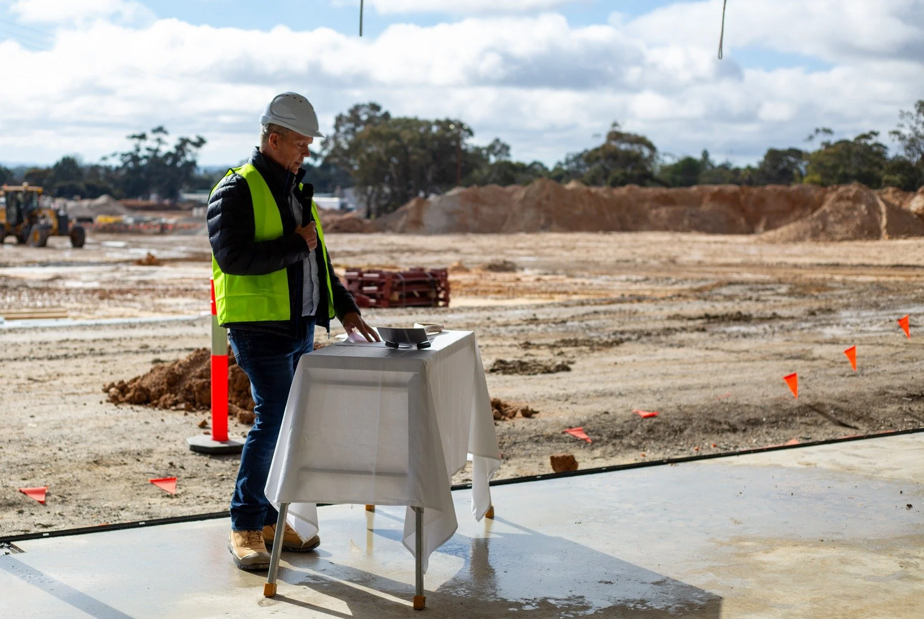 Jamie, Director, wearing a white hard hat and high-visibility vest, standing next to a table with papers on it, at the Nuriootpa Traders construction site with excavated land and construction equipment in the background.