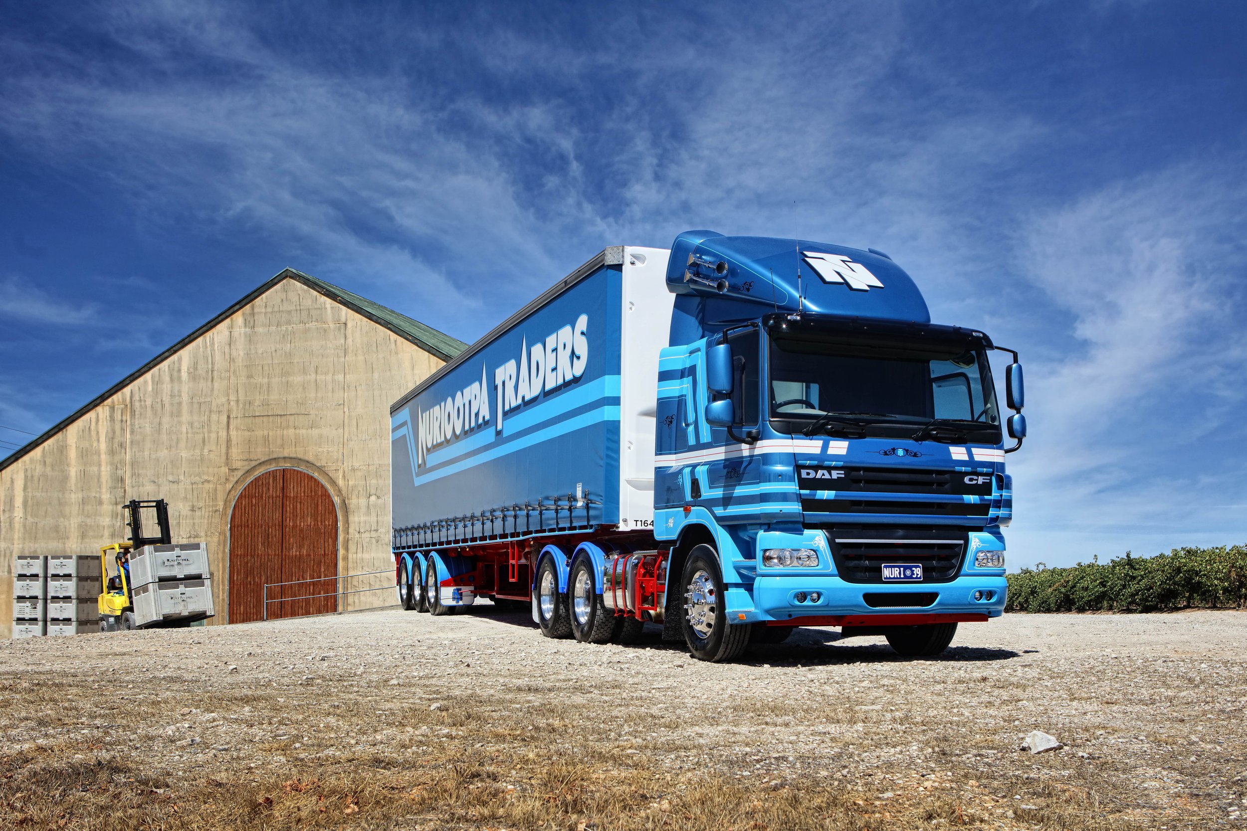 Blue truck with 'NURIOOTPA TRADERS' logo parked near a beige building with a green roof and large wooden doors, under a partly cloudy sky.
