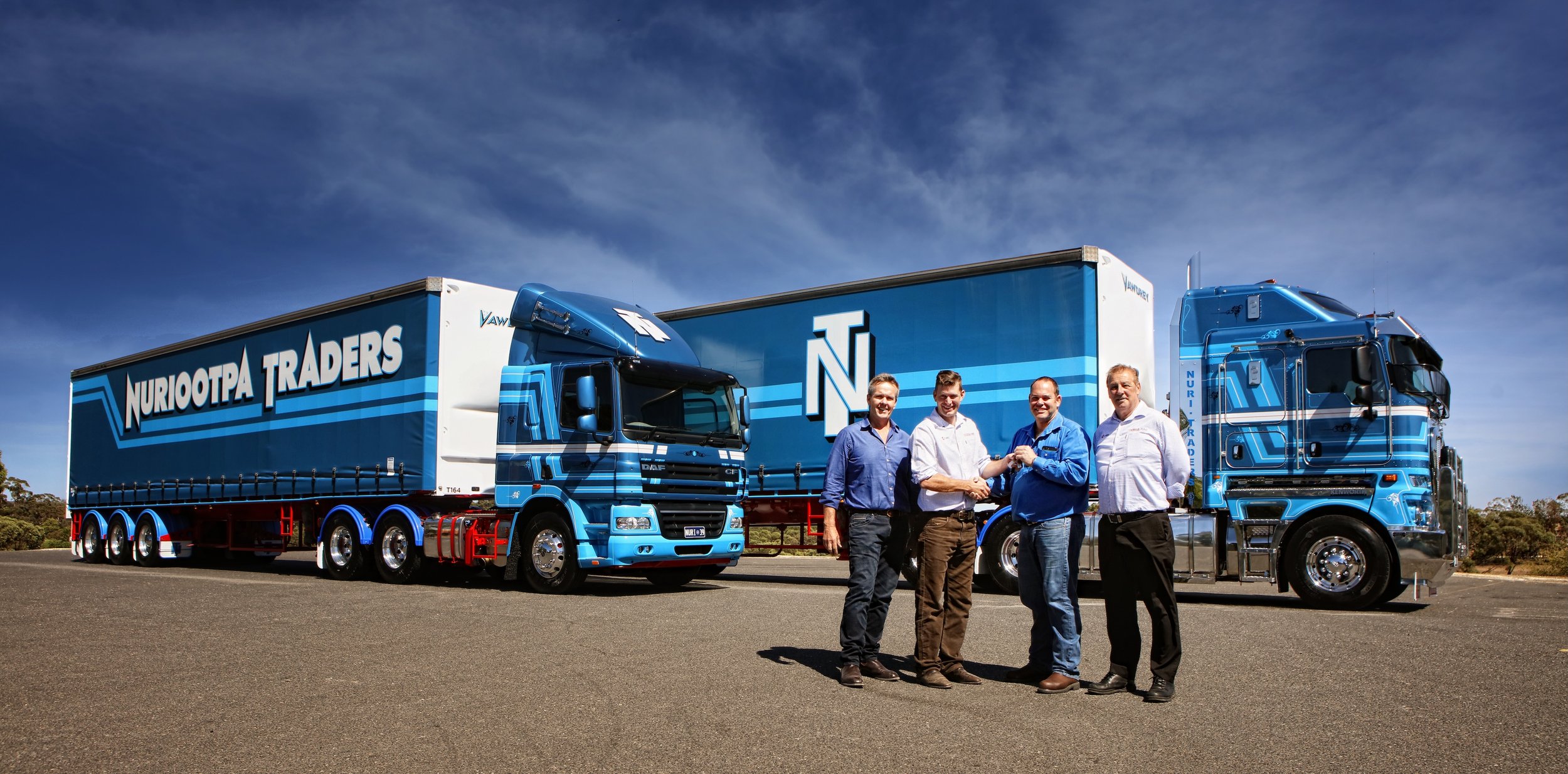 Four men in business attire shaking hands in front of a large, Nuriootpa Traders truck, parked on an open road under a blue sky.