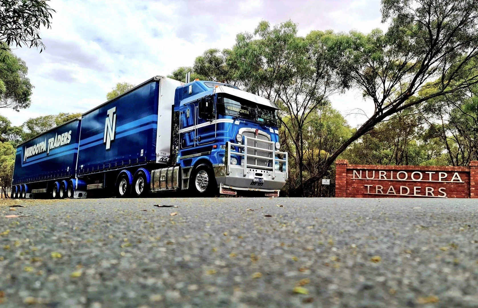 A Nuriootpa Traders semi-truck with white and silver accents parked on the side of a road next to a brick sign. There are green trees and a cloudy sky in the background.