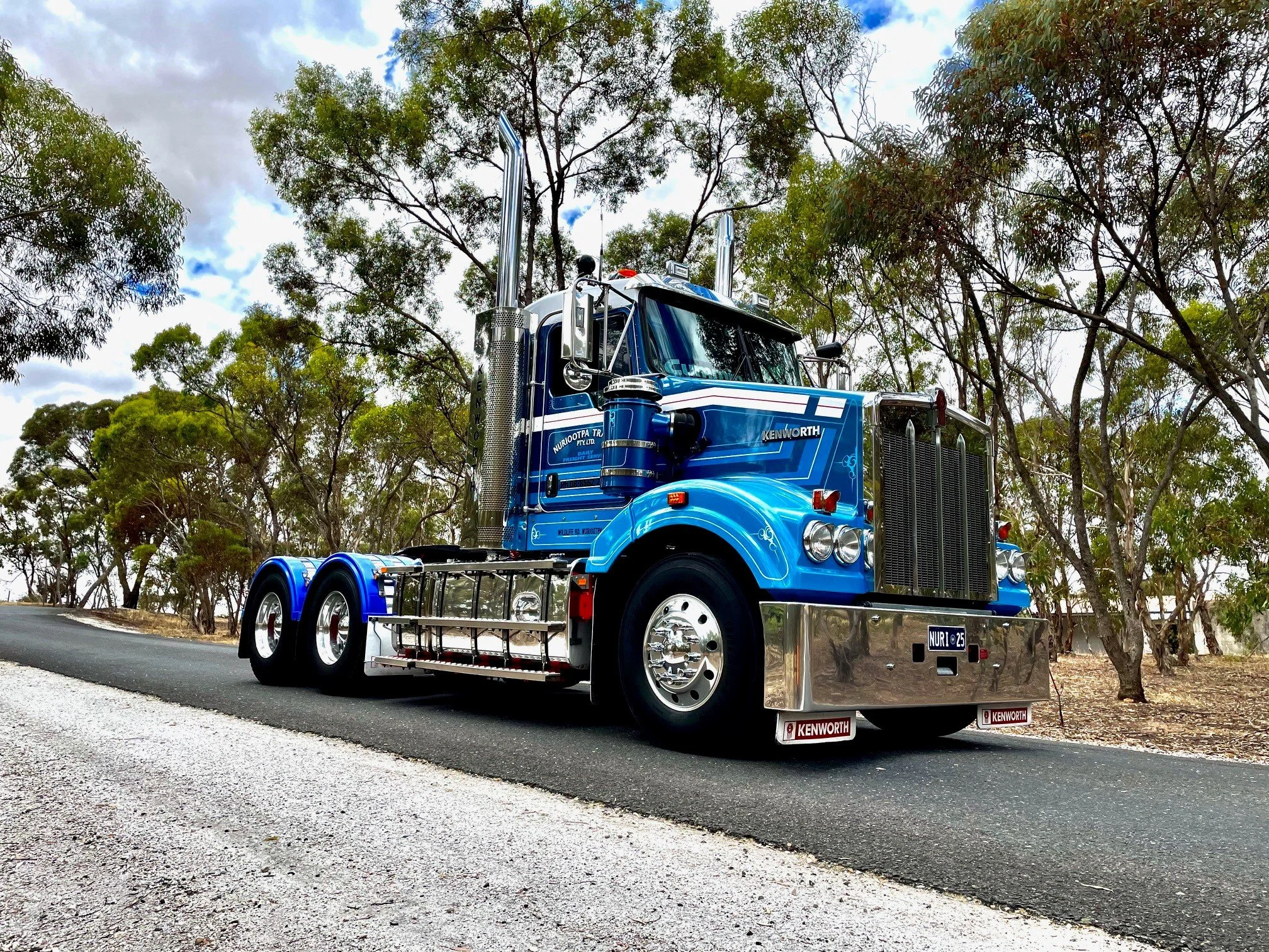 A Nuriootpa Traders Kenworth semi-truck parked on the side of a road with trees and a cloudy sky in the background.