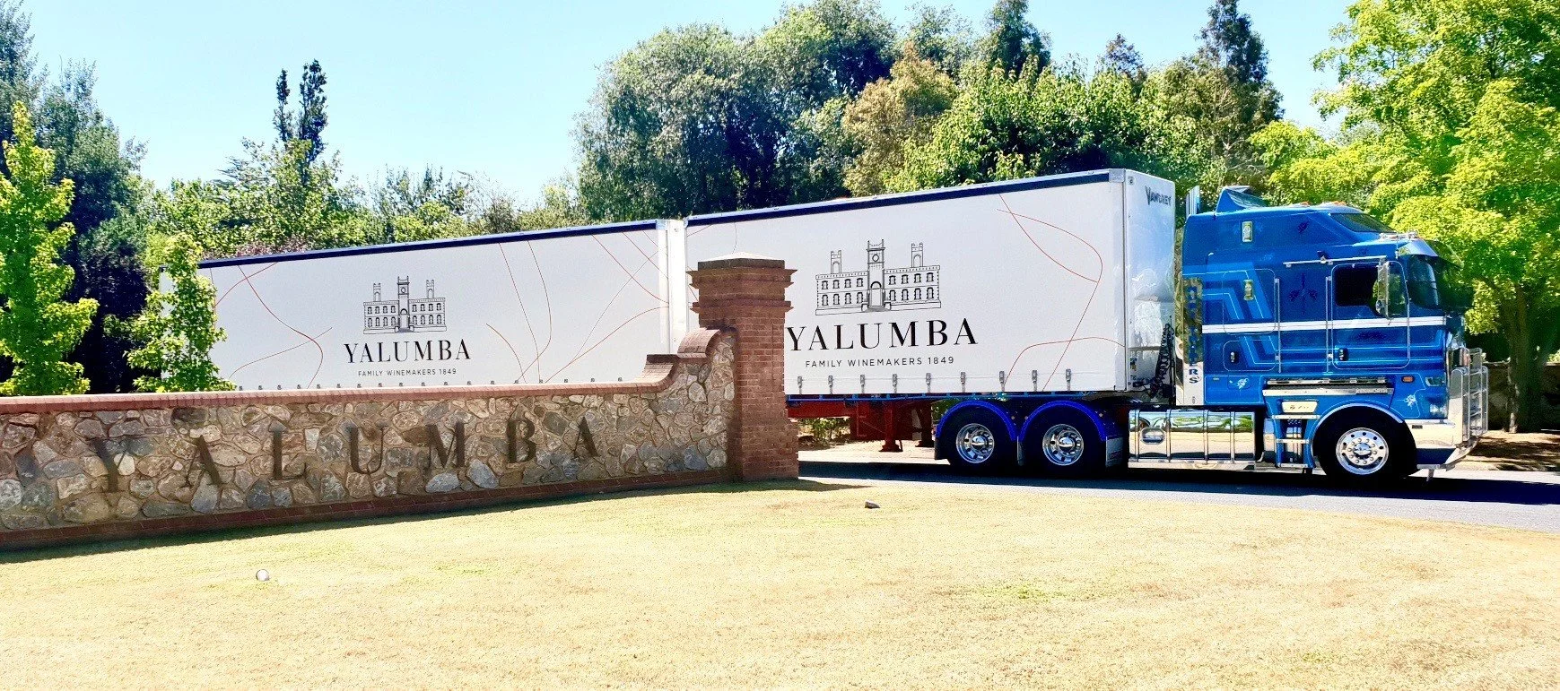 A large blue and white semi-truck with Yalumba branding on the trailer is parked next to a stone and brick wall that has 'YALUMBA' engraved on it. Green trees and a clear blue sky are in the background.