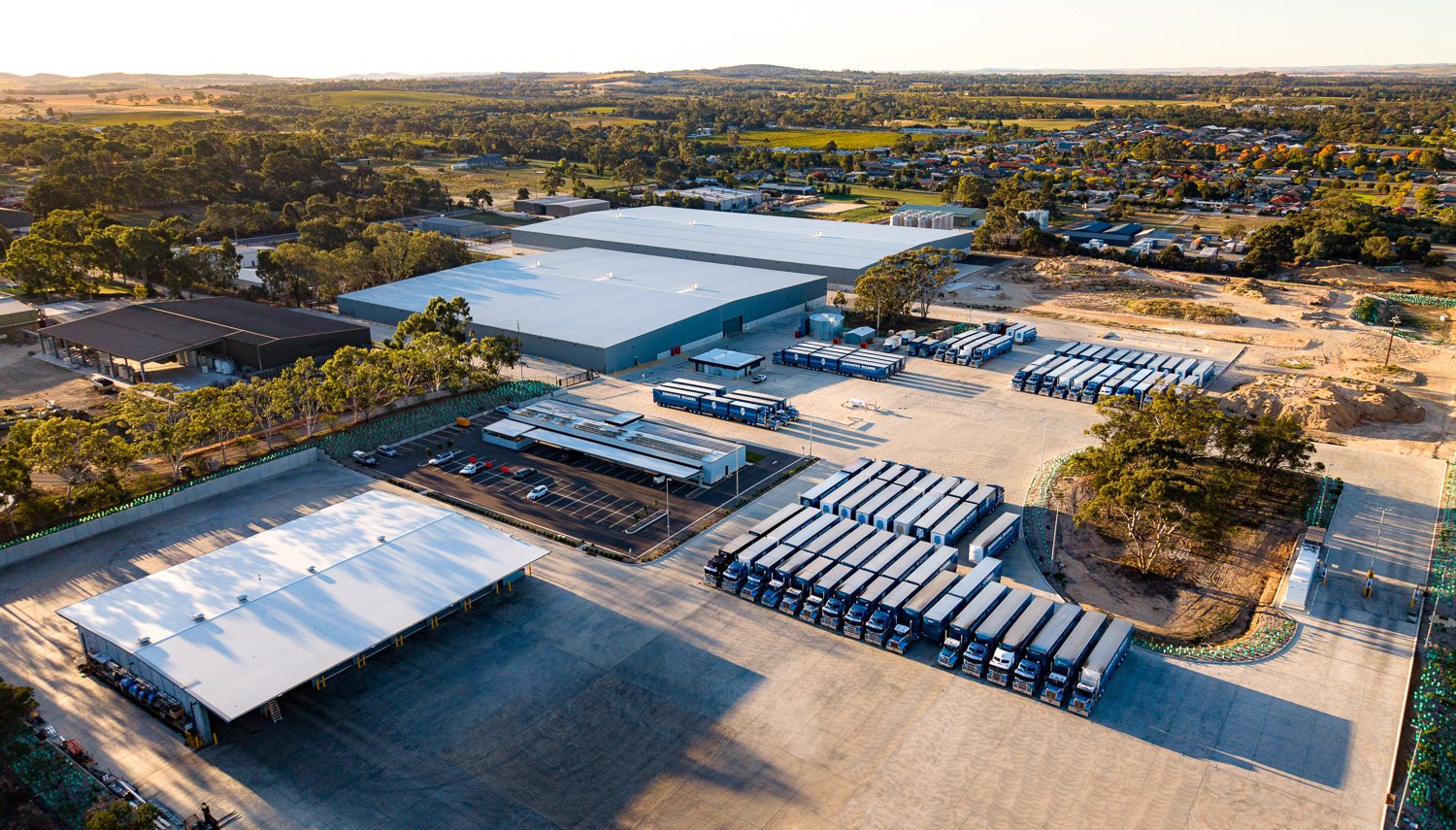 Aerial view of Nuriootpa Traders in the Barossa Valley, with several sections, parking lots, and a fleet of trucks parked in designated areas, surrounded by greenery and open land.