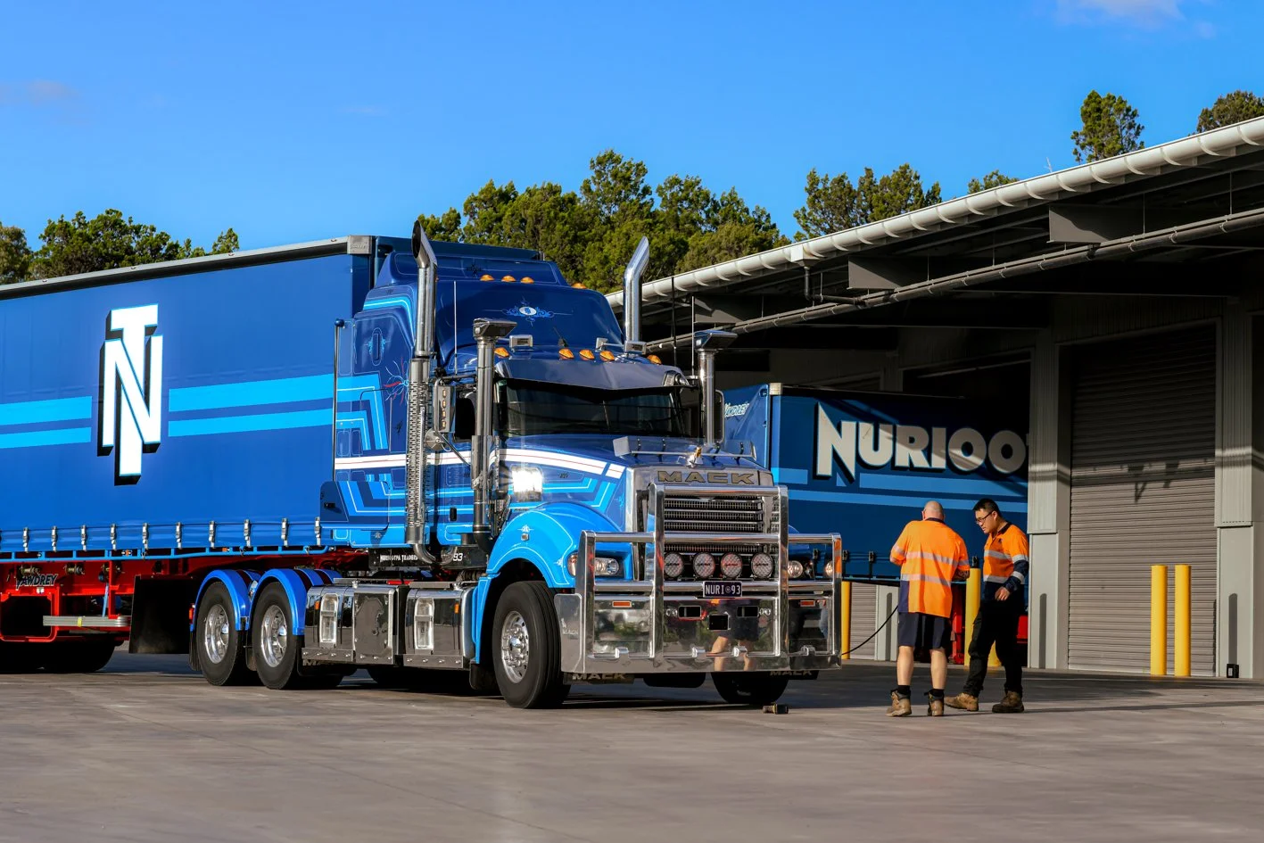 Nuriootpa Traders semi-truck parked outside a warehouse with two workers in orange safety vests near the truck.
