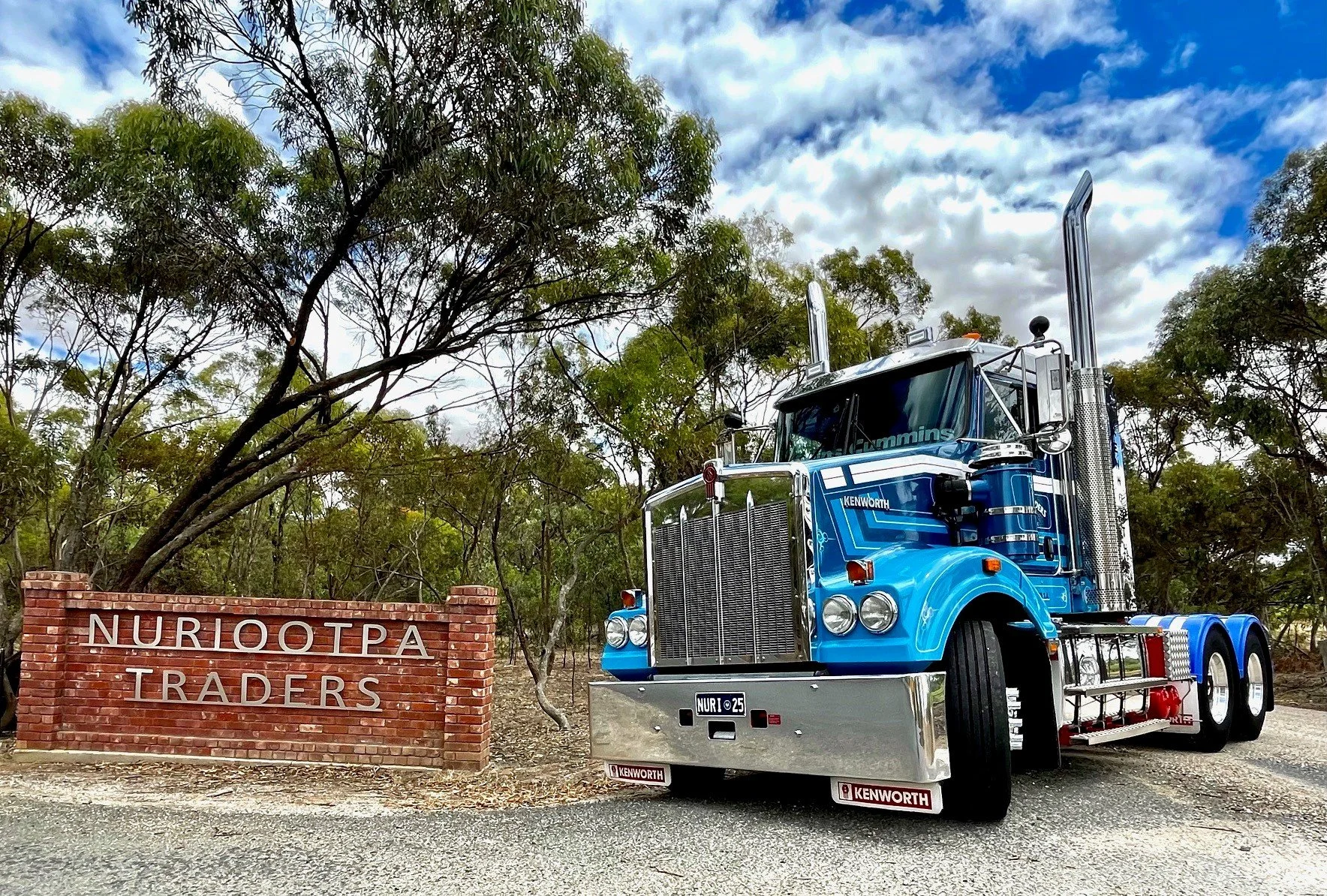 Nuriootpa Traders Kenworth semi-truck parked near a brick sign that reads "NURIOOTPA TRADERS." The truck is in a natural setting with trees and a partly cloudy sky in the background.