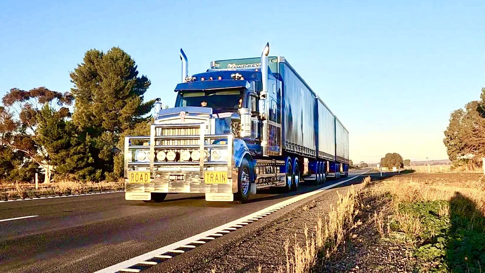 Nuriootpa Traders semi-truck with a trailer driving on a rural highway in the late afternoon, surrounded by trees and dry grass.