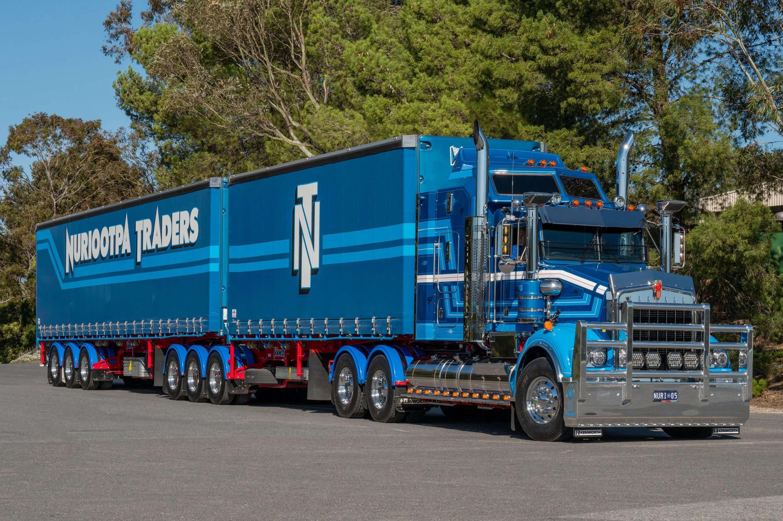 A Nuriotpa Traders truck parked outdoors on a paved surface, with tall green trees and a clear blue sky in the background.
