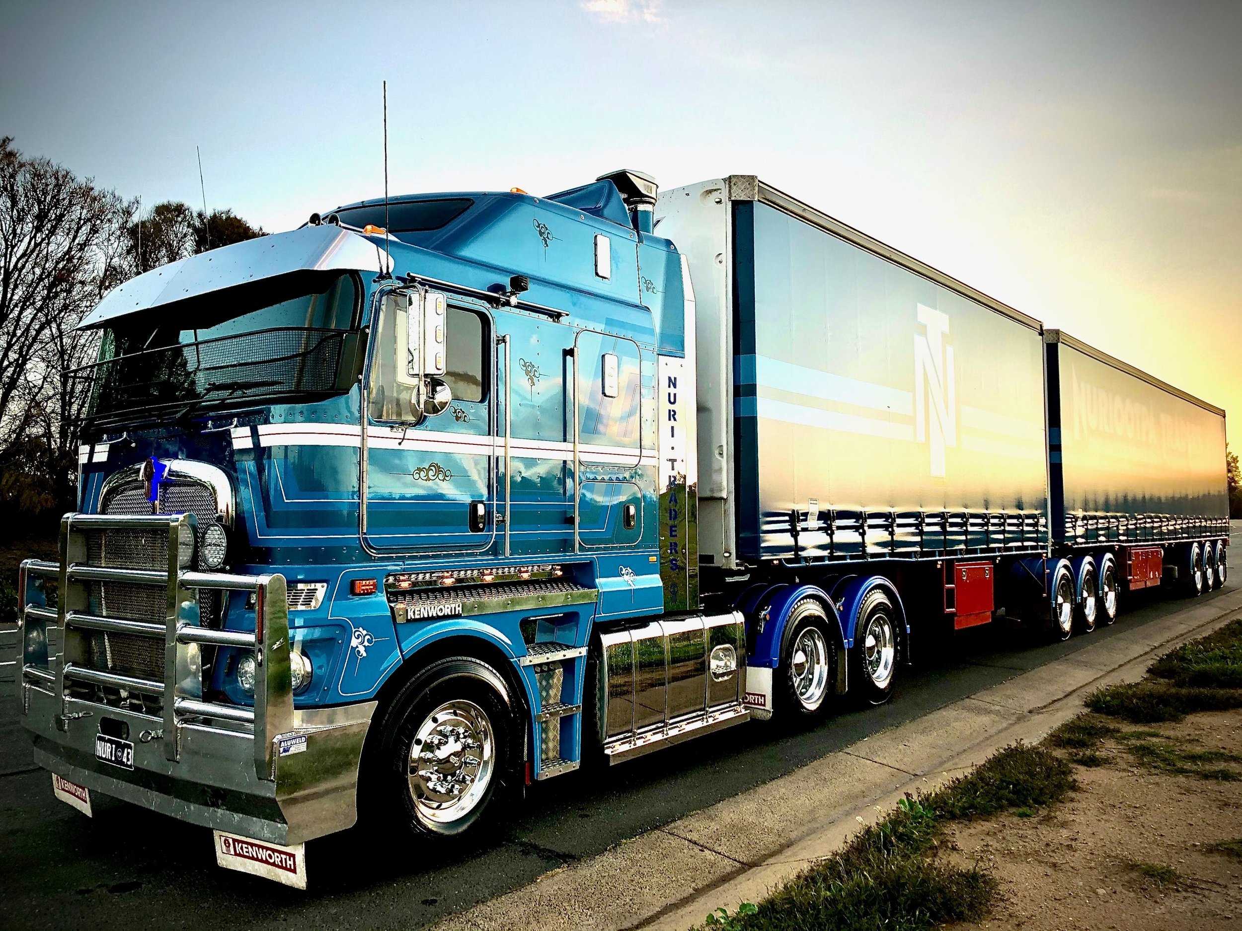 A Nuriootpa Traders semi-truck with decorative details parked on a street at sunset, with trees in the background.