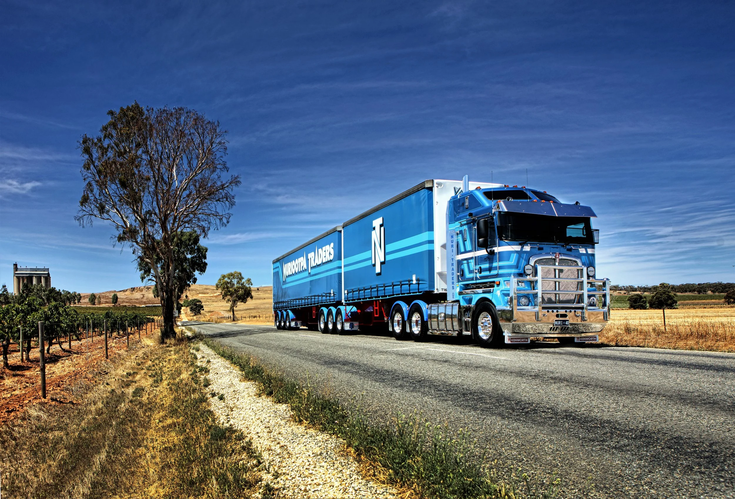 A Nuriootpa Traders truck driving on a rural road with a tree and fields in the background under a partly cloudy sky.