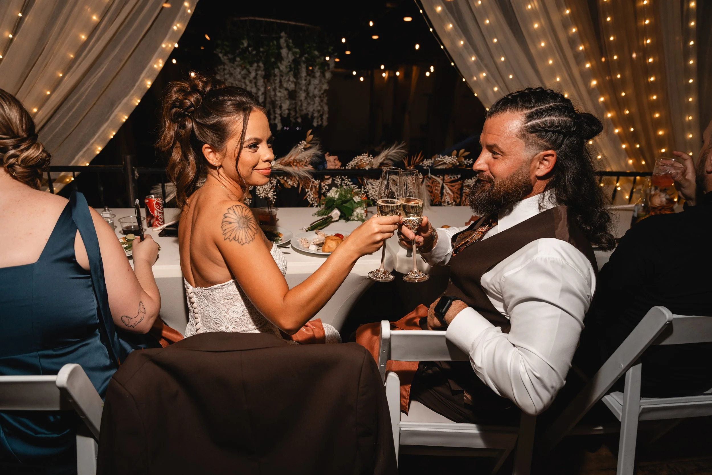 A romantic couple celebrating with champagne at a decorated wedding reception, toasting while sitting at a banquet table with guests and festive lights in the background.