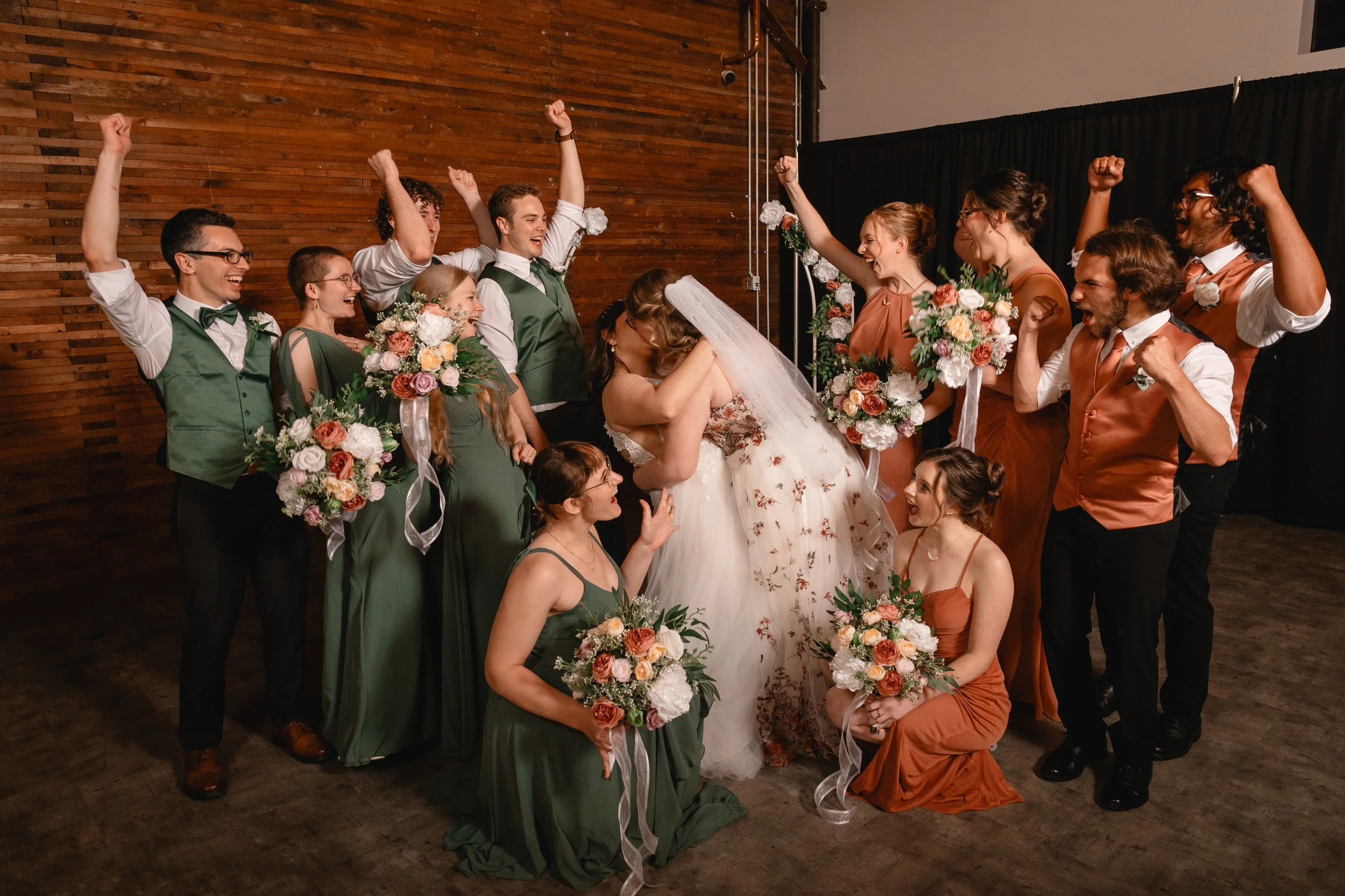 A bride and groom kissing surrounded by their wedding party, with friends and family celebrating, holding bouquets, and raising their fists in excitement indoors with a brown wooden wall background.