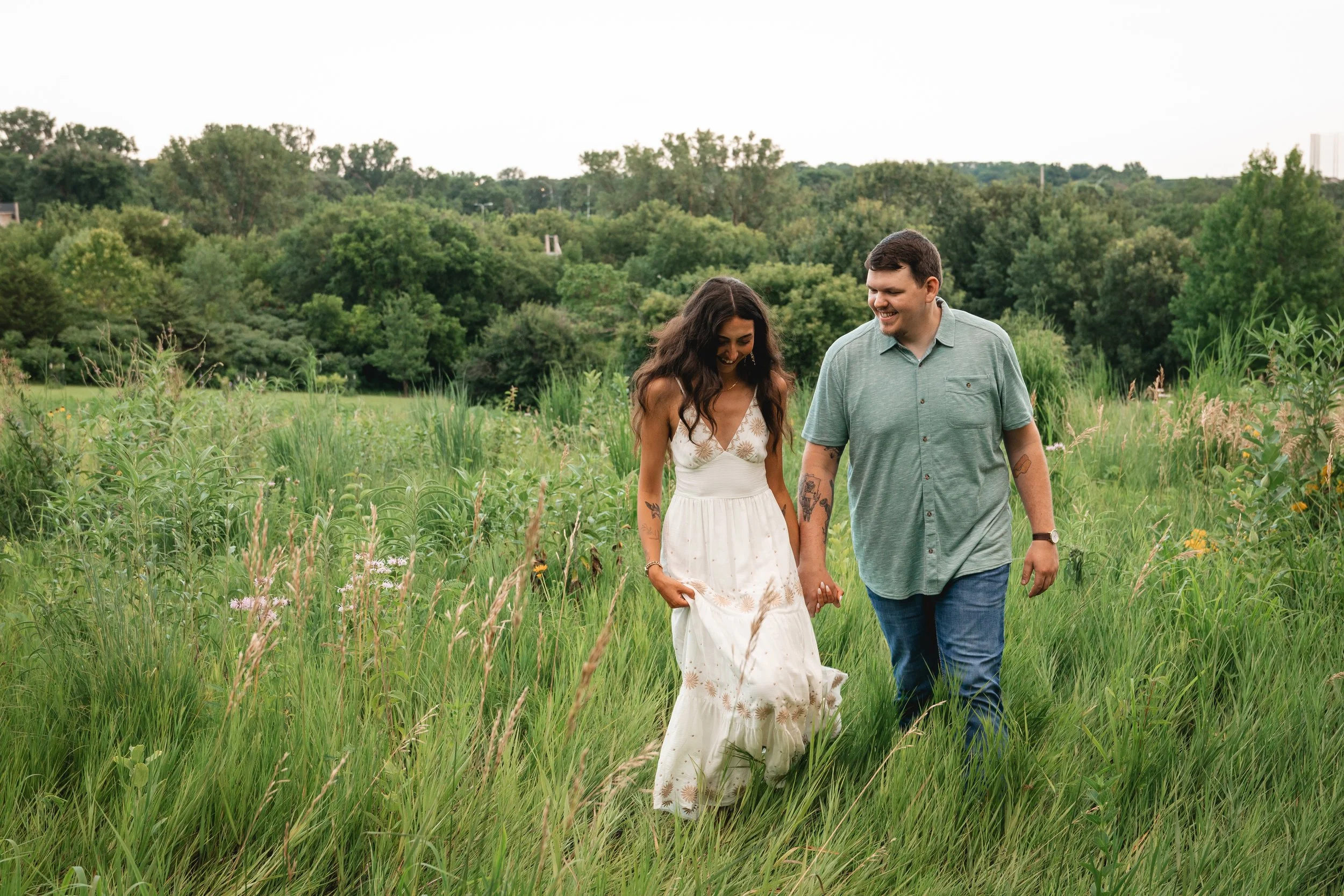 A couple holding hands and walking through a grassy field with lush green trees in the background, smiling and enjoying each other's company.