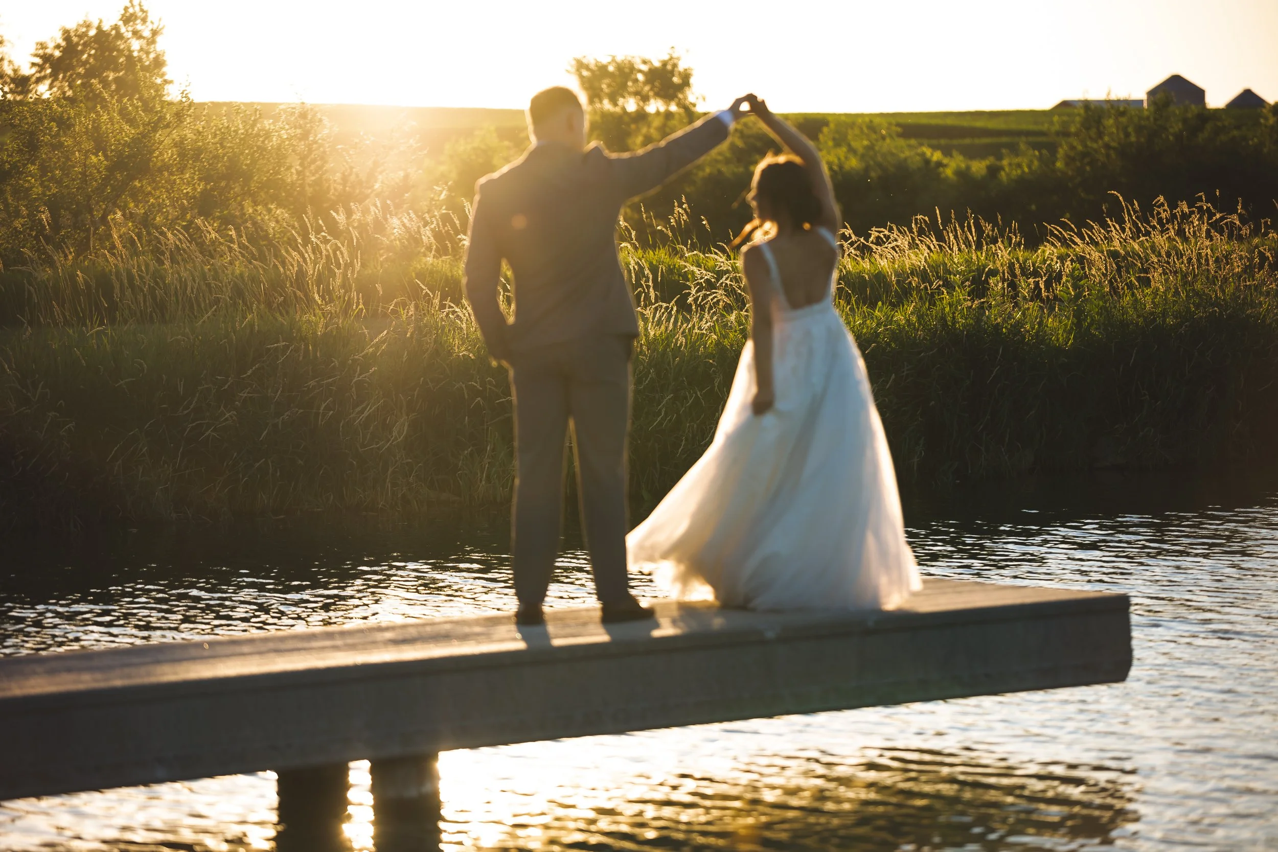 A couple in wedding attire dancing on a dock by a lake during sunset.