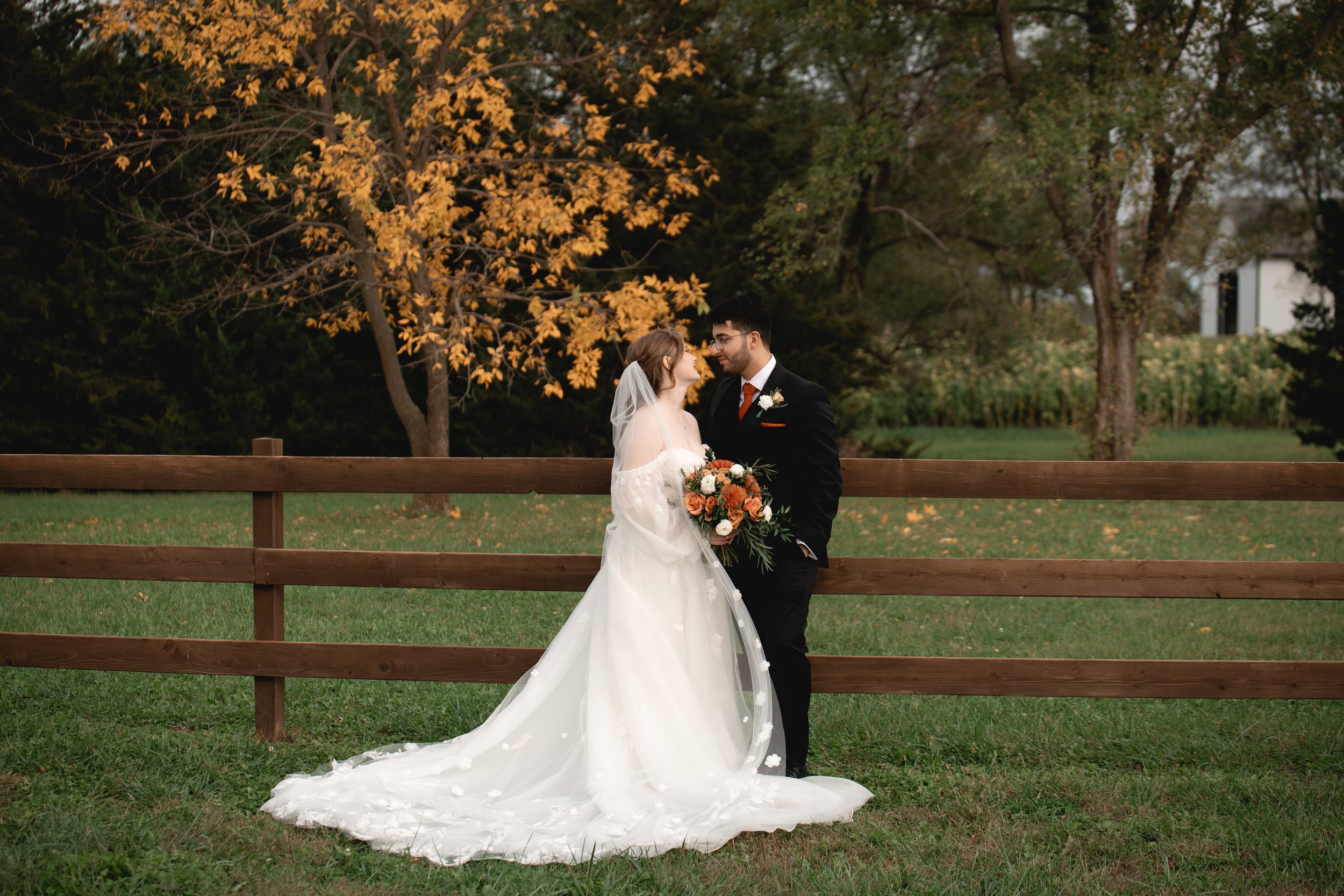 A bride in a white wedding gown with a long train and veil, holding a bouquet of flowers, and a groom in a black suit with a white shirt and orange tie, standing close together outdoors behind a wooden fence with trees and autumn leaves in the background.