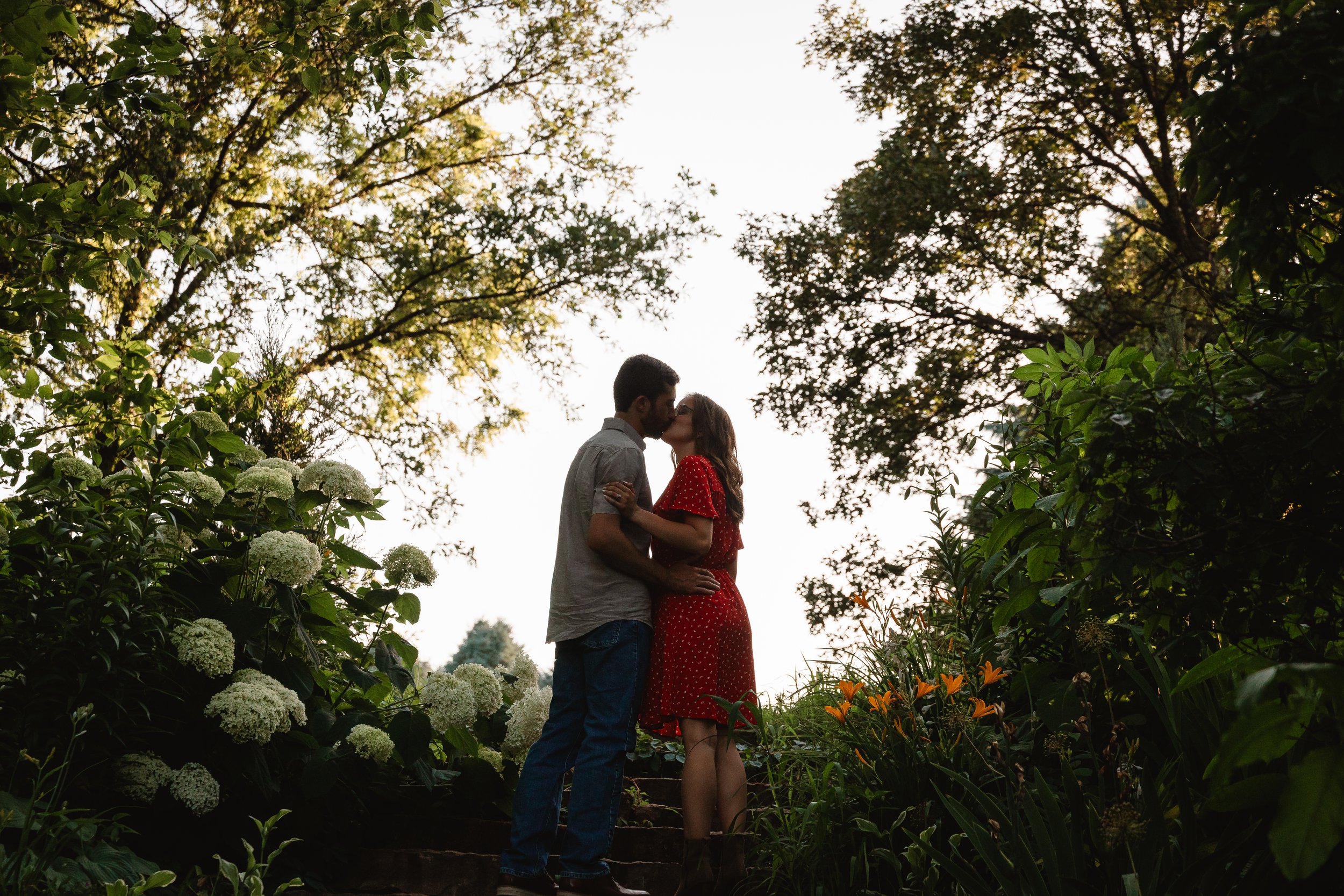 A couple sharing a kiss in a garden during sunset, surrounded by green foliage and white and orange flowers.