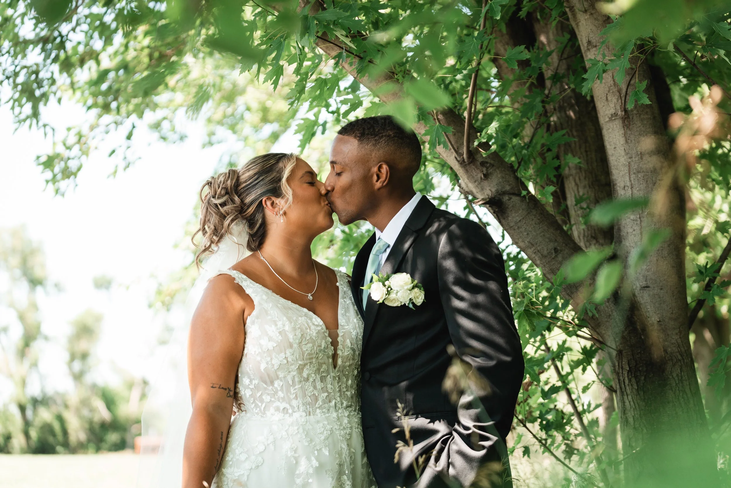 A bride and groom kissing outdoors beneath a tree with green leaves, during their wedding. The bride is wearing a white lace dress and necklace, and the groom is in a black suit and tie with a white boutonniere.