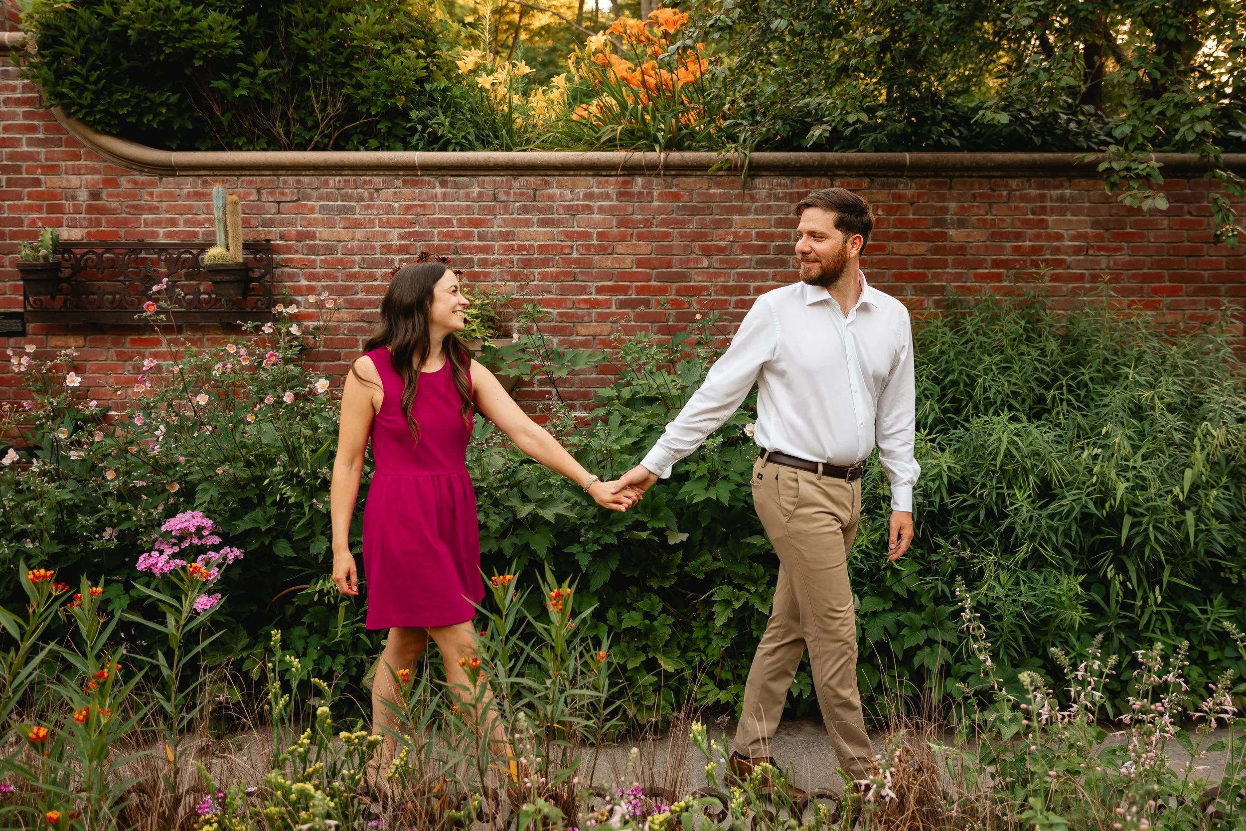 A couple walking hand in hand through a garden with various flowers and plants, with a red brick wall in the background.