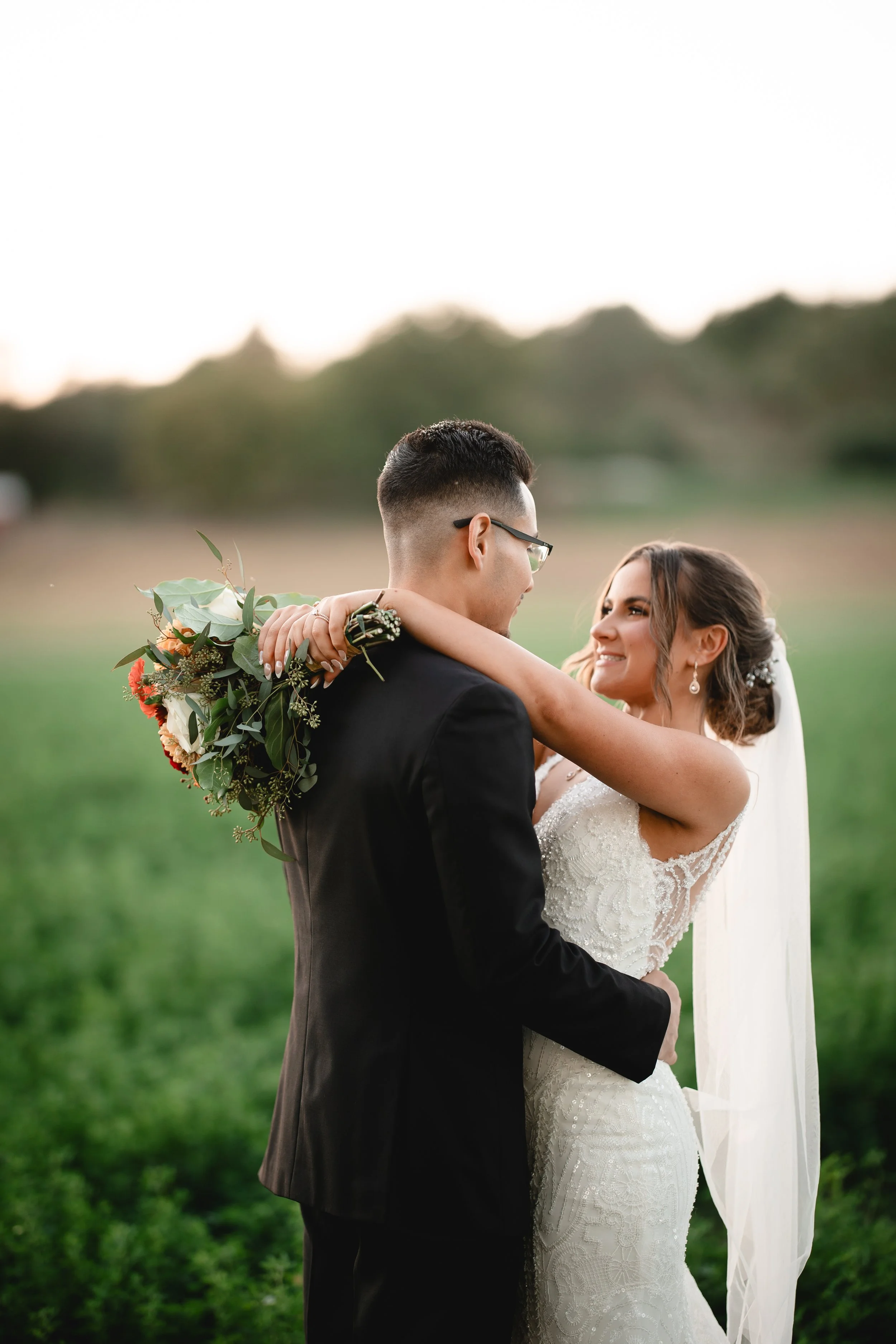 A bride and groom embracing outdoors during their wedding, smiling at each other, with greenery and trees in the background.
