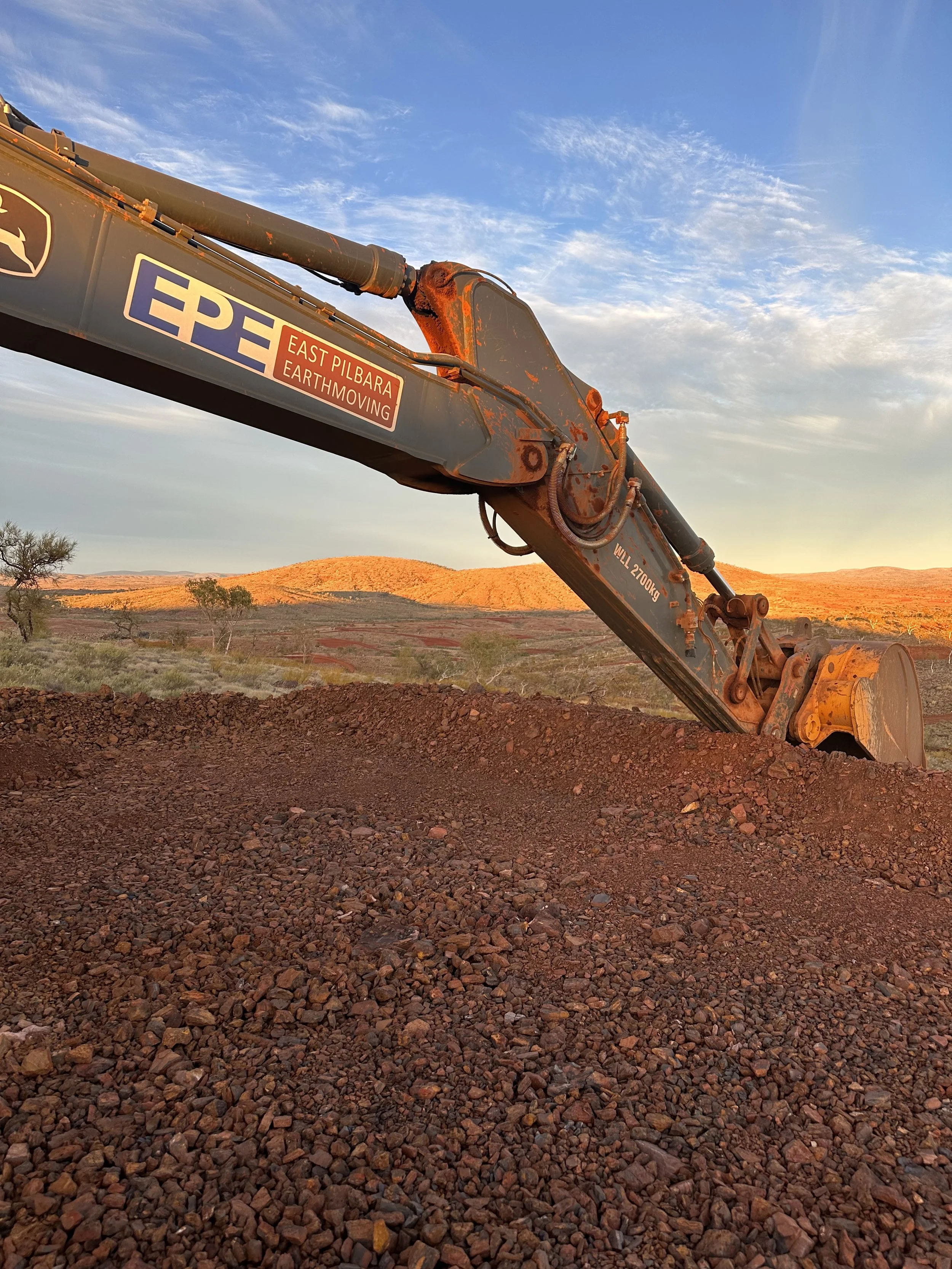 Close-up of a large earth-moving excavator arm with a soil pile in a landscape with hills and a partly cloudy sky at sunset.