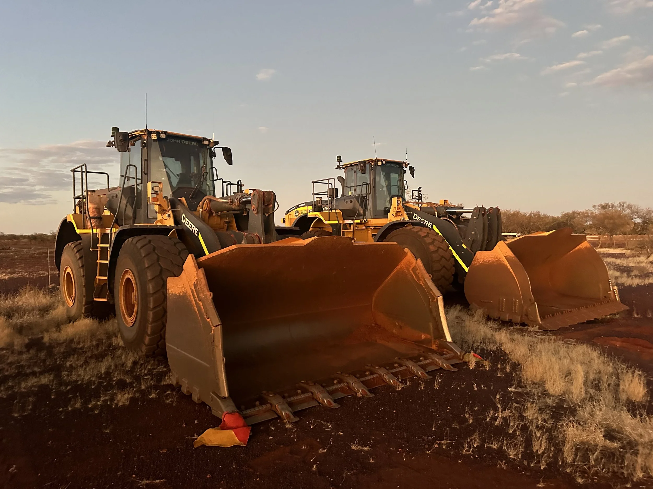 Two large yellow and black John Deere bulldozers working on soil in an open field during sunset.