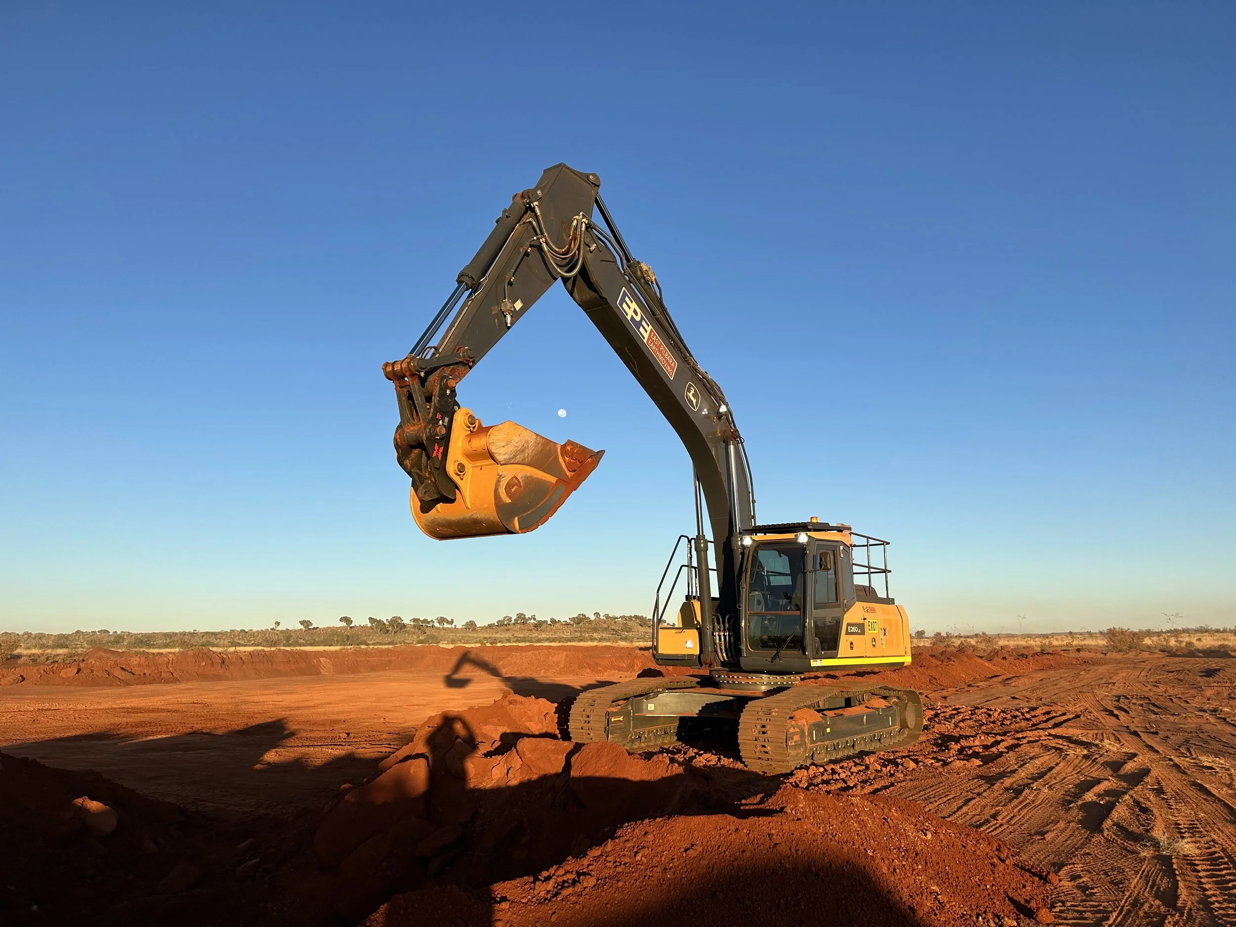 An excavator with a yellow bucket working on reddish soil in a field, under a clear blue sky with a small moon visible near the horizon.