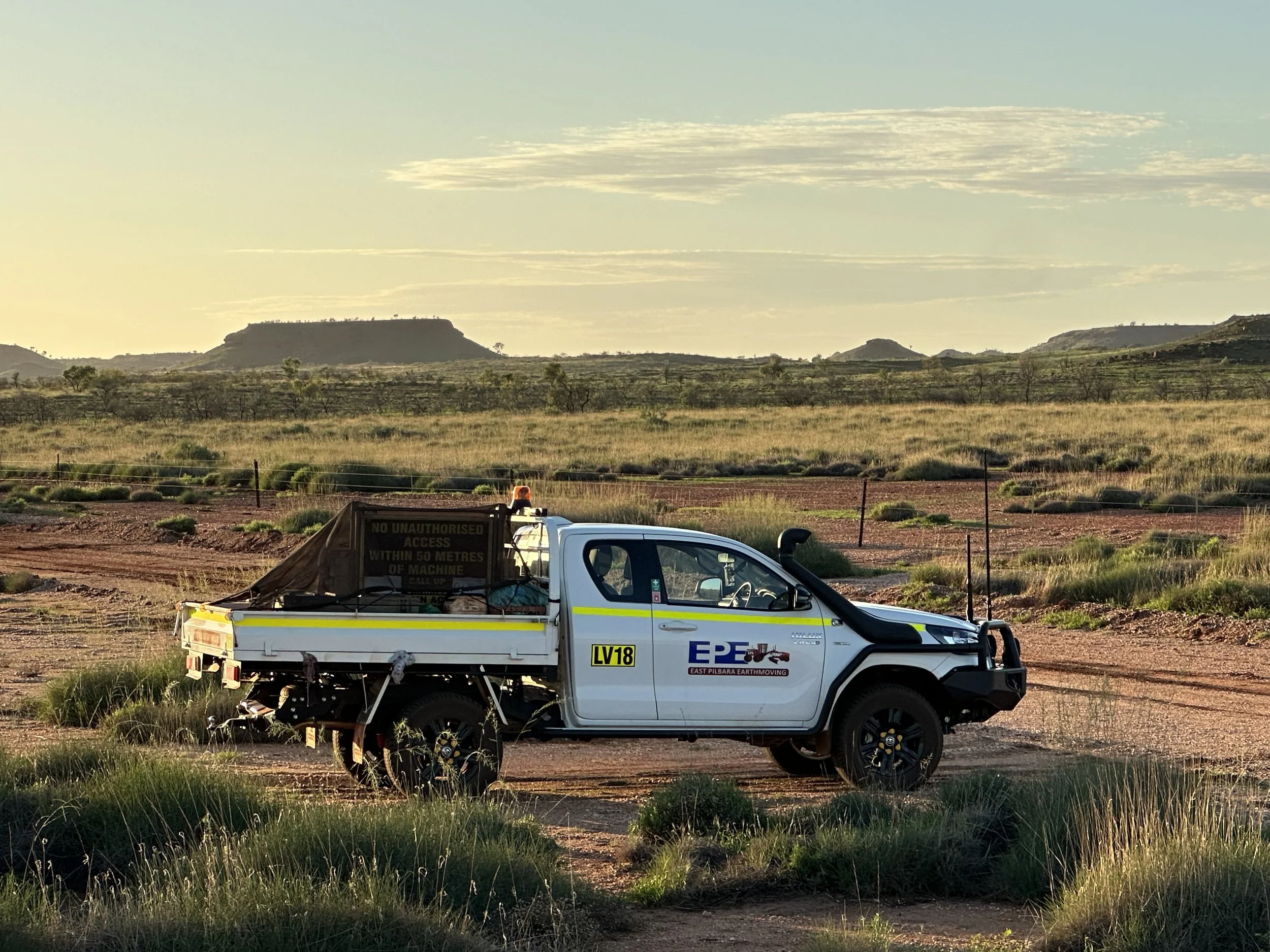 A white utility vehicle with yellow and black markings, parked on a dirt area near a grassy landscape with small trees. In the background, there are rolling hills and a clear sky with some clouds.
