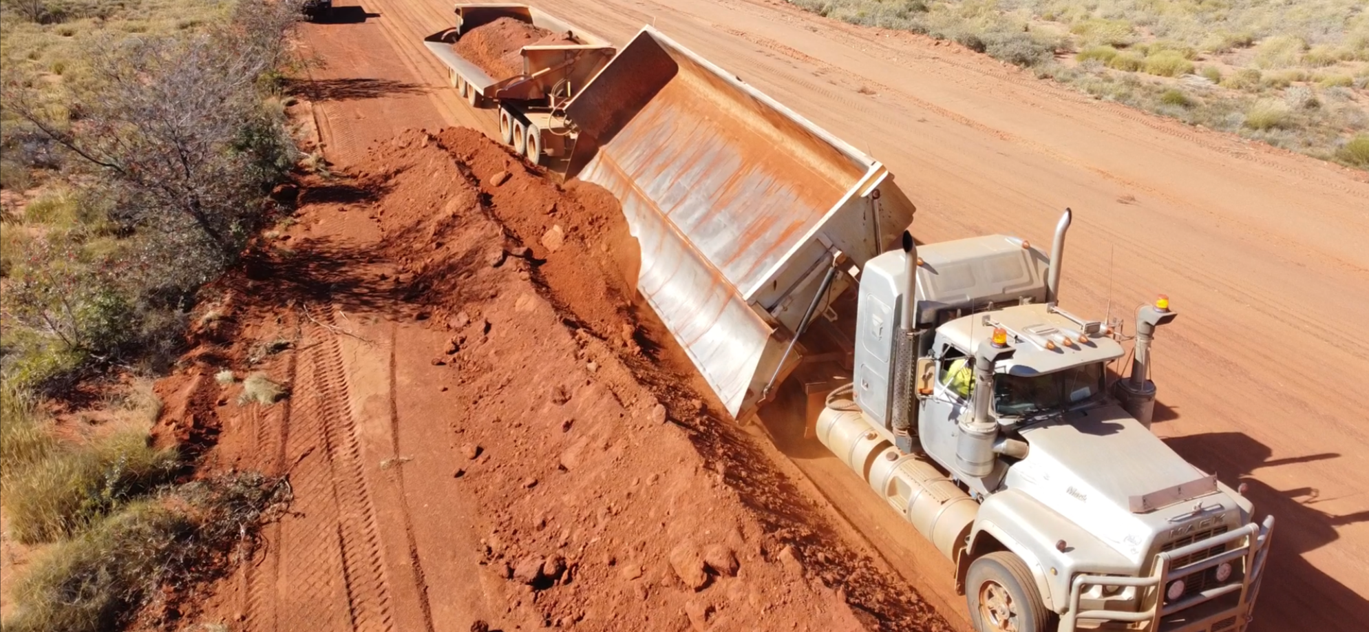 A dump truck is unloading red dirt onto a dirt road in a rural, desert-like area with sparse vegetation.
