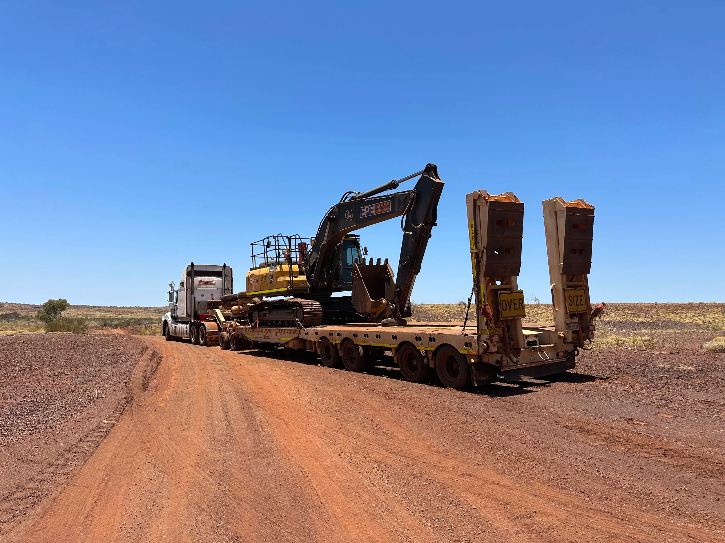 A large truck transporting heavy construction equipment including a John Deere excavator on a flatbed trailer on a dirt road in a dry, open landscape.