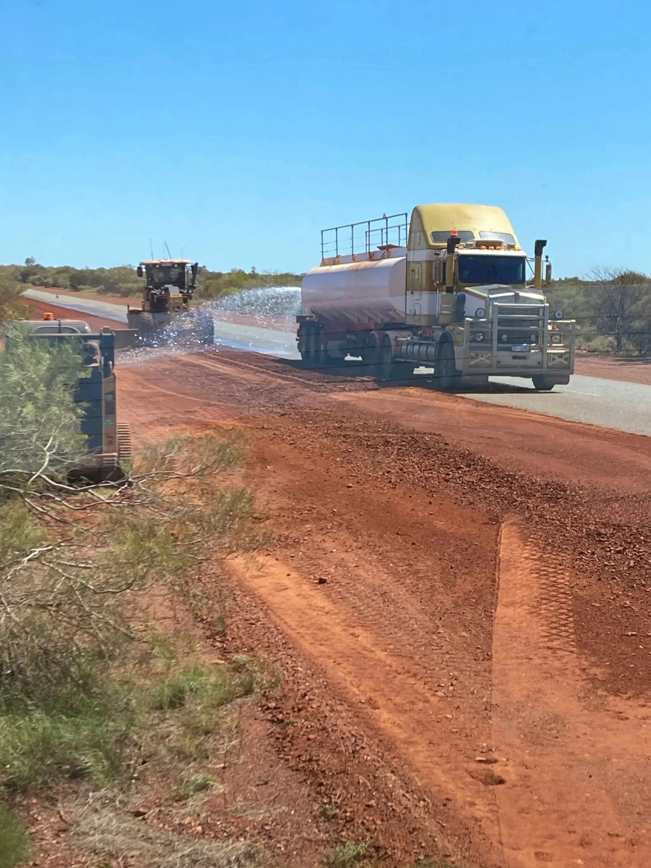 Two large construction trucks working on a dirt road in a rural area under a clear blue sky.