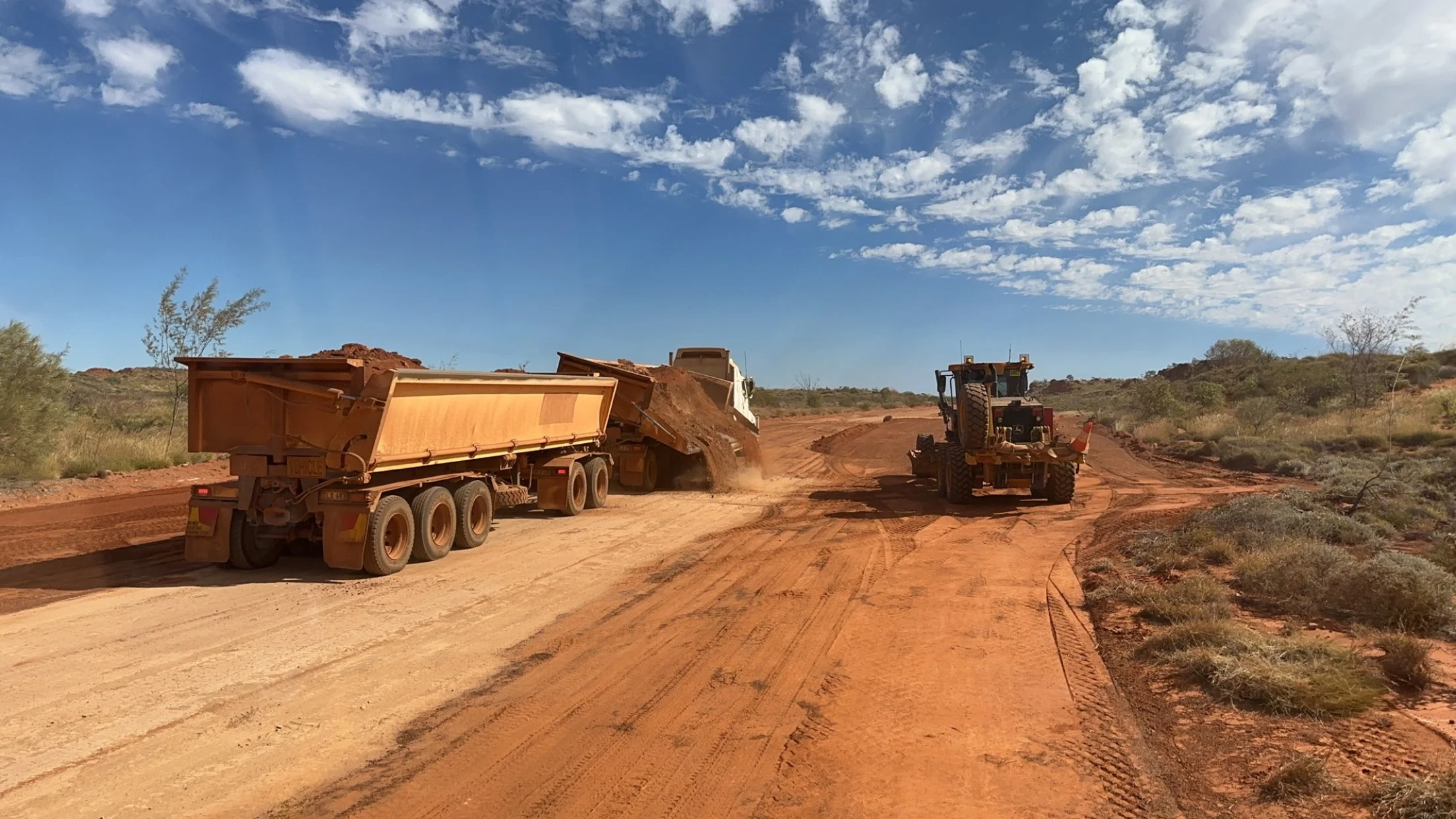 Construction vehicles on a dirt road, with a dump truck spreading dirt and a grader leveling the ground under a blue sky with scattered clouds.