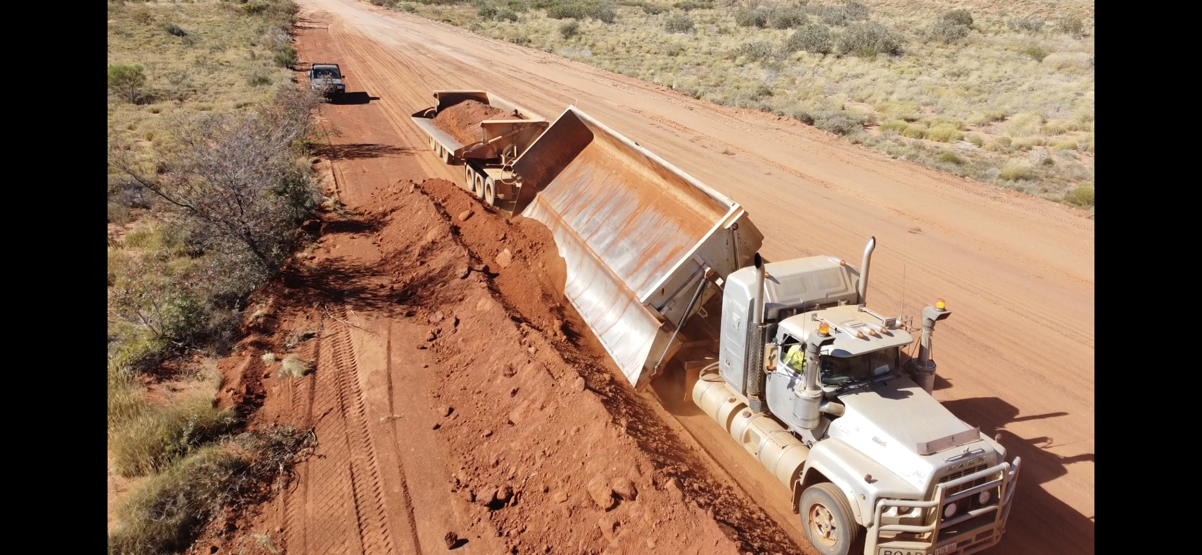 A large dump truck unloading soil on a dirt road in a rural or desert area, with sparse vegetation and an empty and dusty landscape.