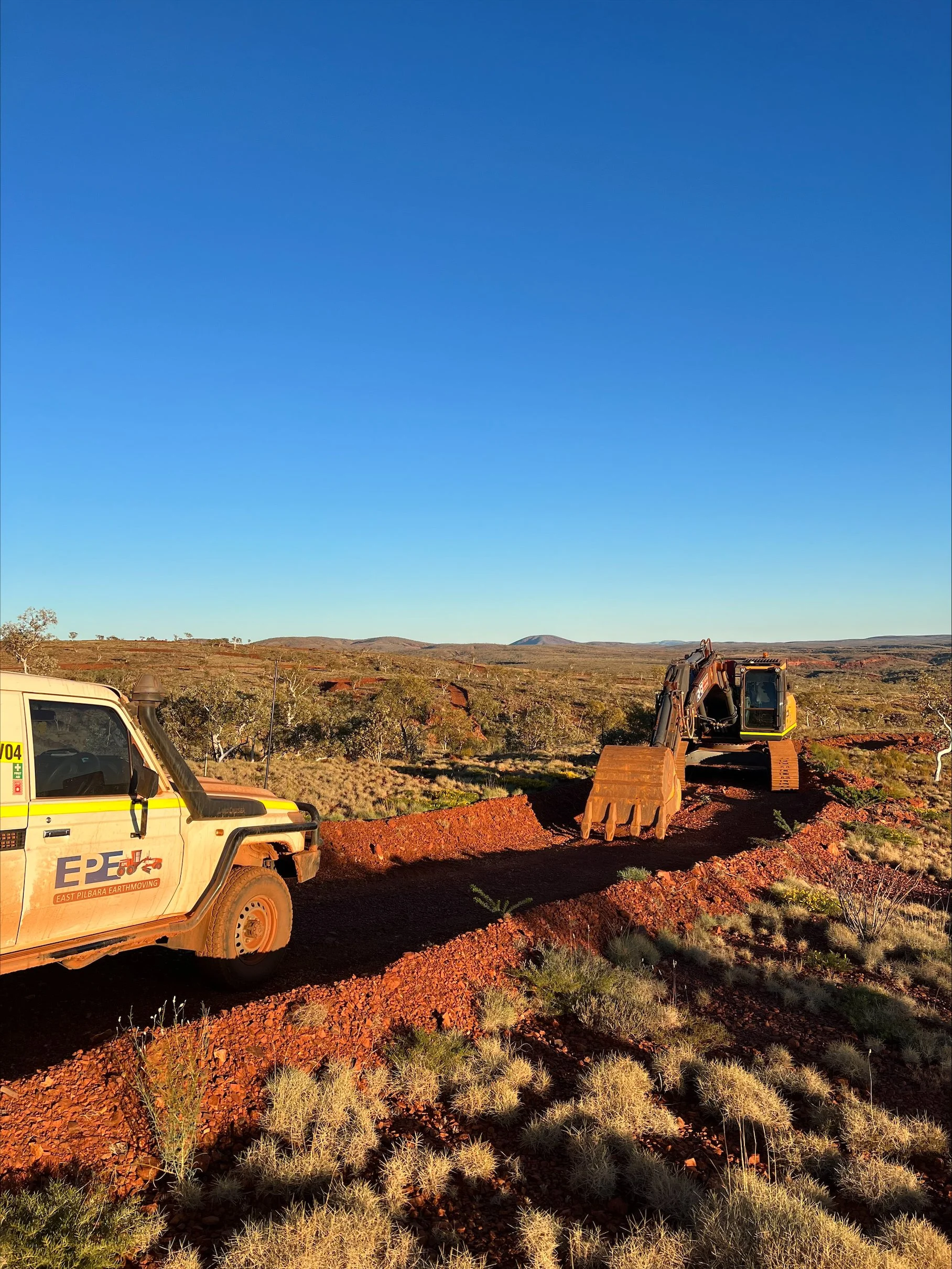 A landscape with a clear blue sky, some green shrubs, and orange-brown earth in the foreground. There are two vehicles: a white truck with EPE Logo on the left side and a yellow excavator on the right side.