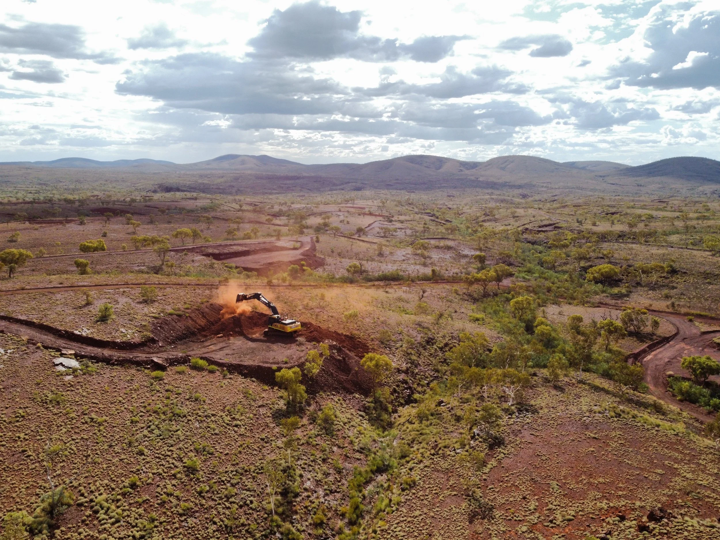 A landscape view of a semi-arid terrain with scattered small trees, mountains in the distance, and a partly cloudy sky. In the foreground, a yellow excavator is digging a trench.