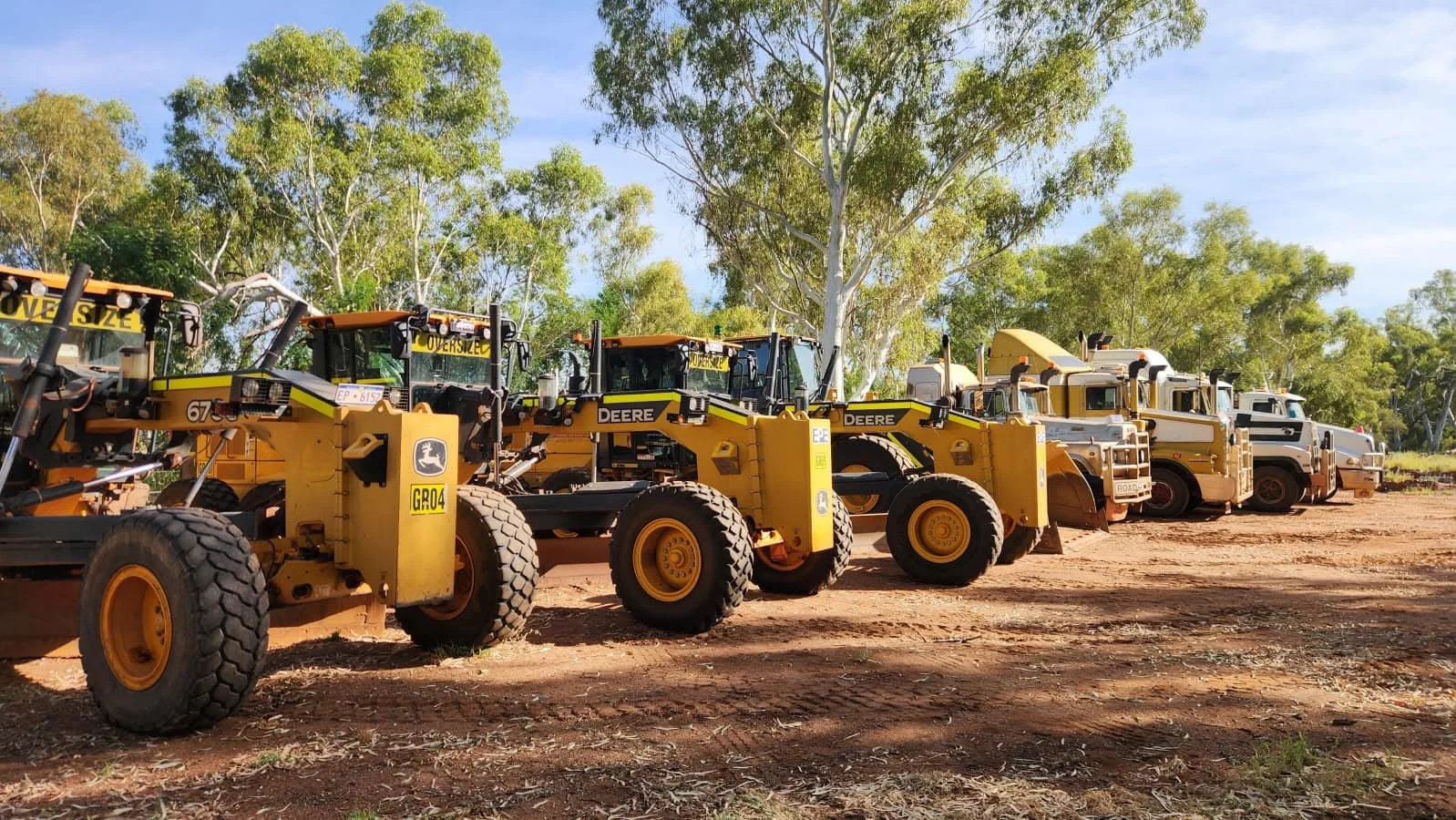 A row of construction vehicles, including bulldozers and dump trucks, parked on a dirt lot under trees.