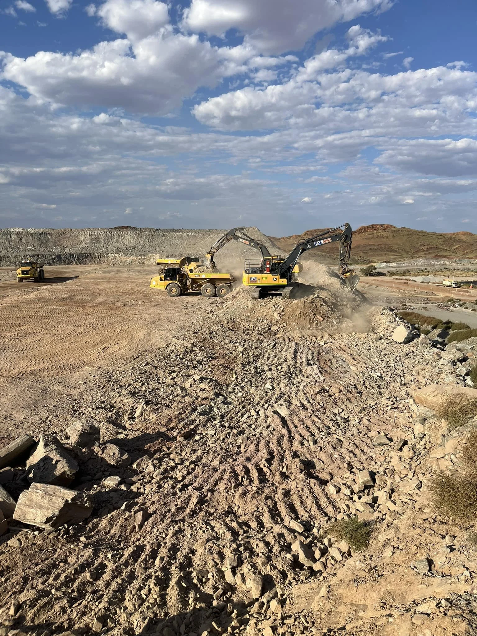 Construction site with yellow excavators moving dirt and rocks under a partly cloudy sky.