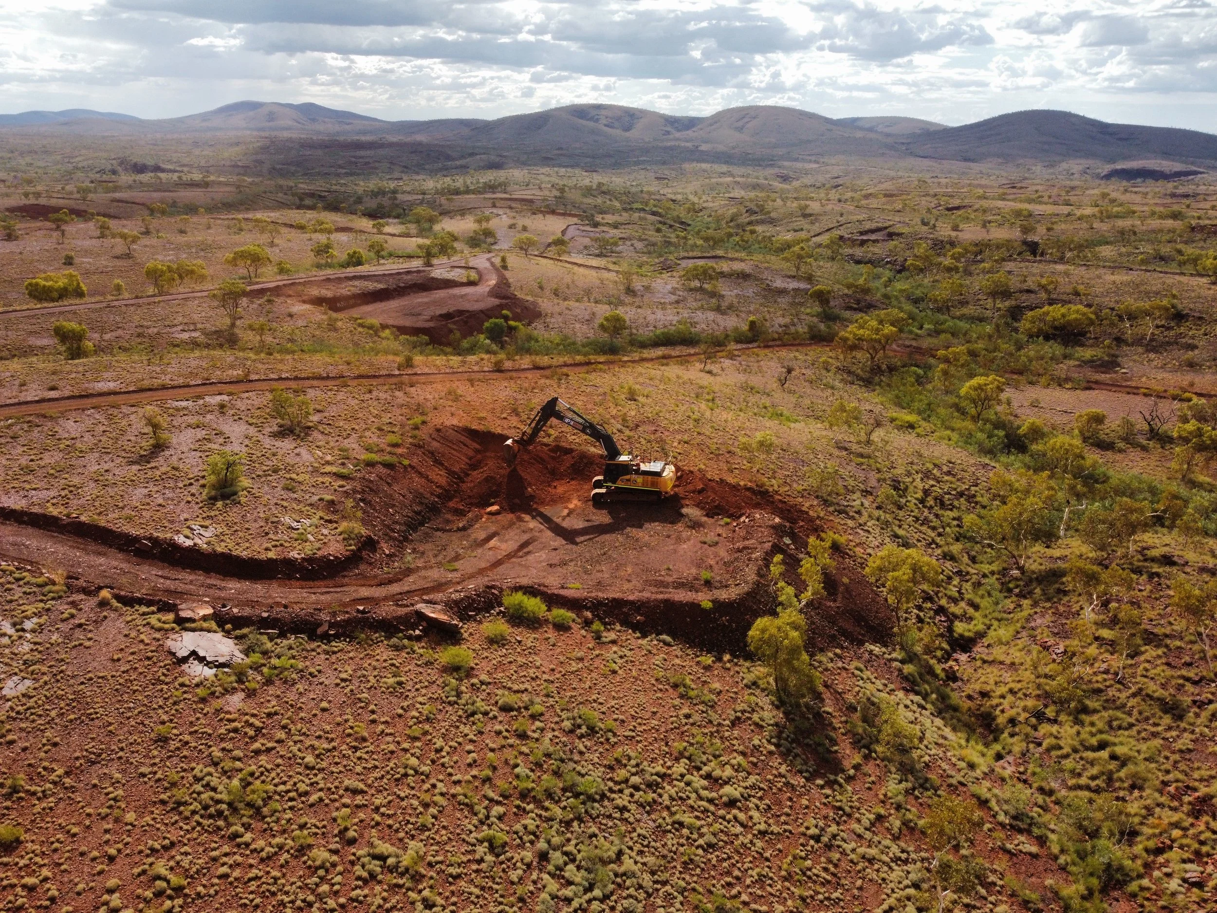 An aerial view of a construction site in a semi-arid landscape, with a small excavator digging a hole and light green trees scattered across the terrain under a cloudy sky.