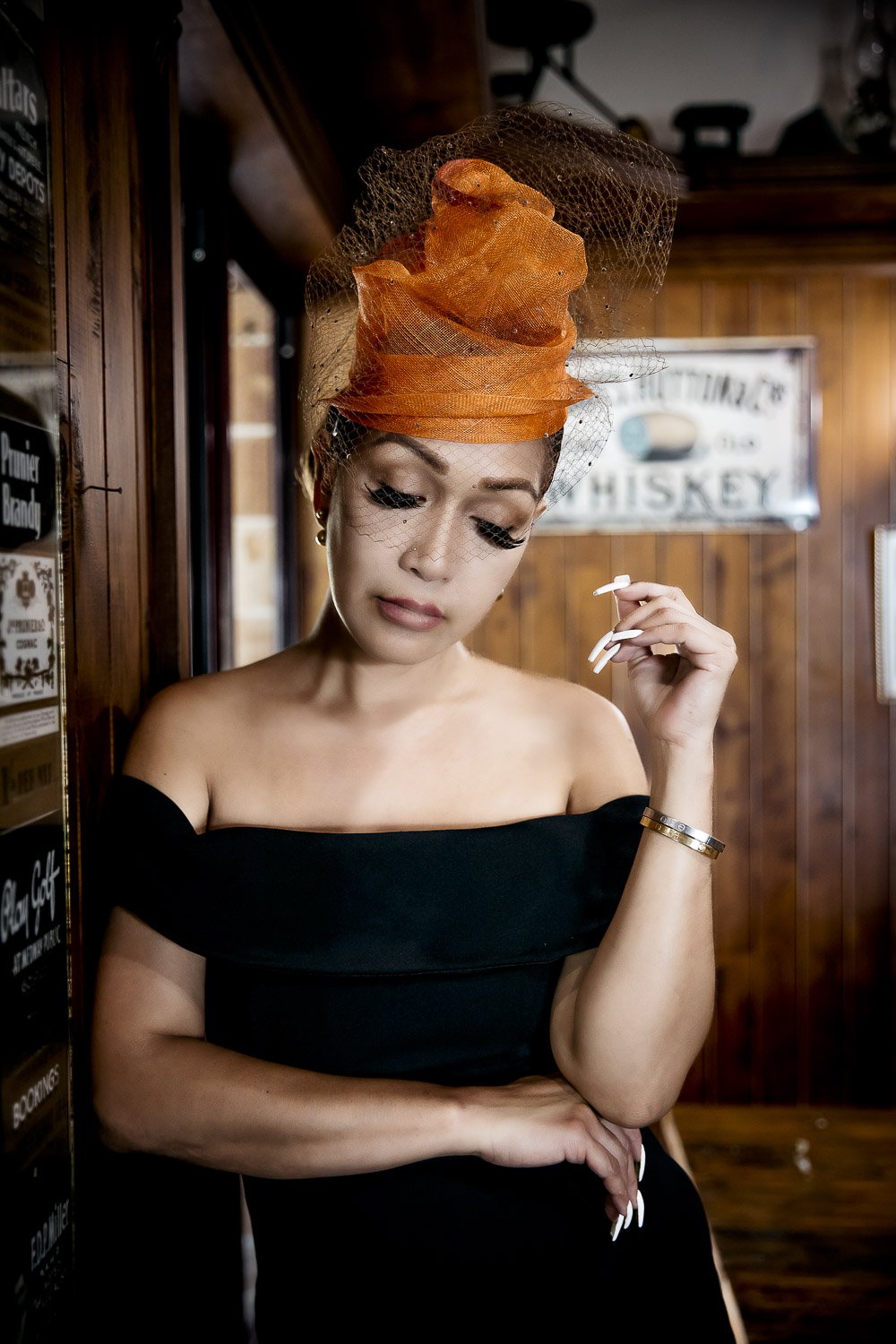 Woman wearing an orange fascinator and black dress, standing indoors with wood paneling background.