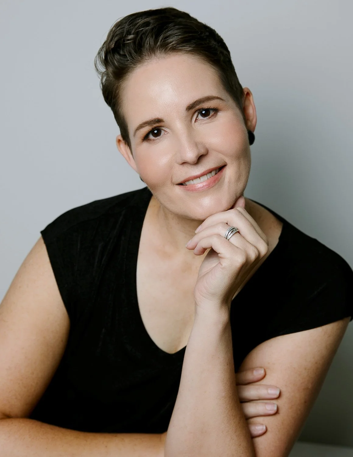 Person with short hair smiling and resting their chin on their hand, wearing a black shirt.