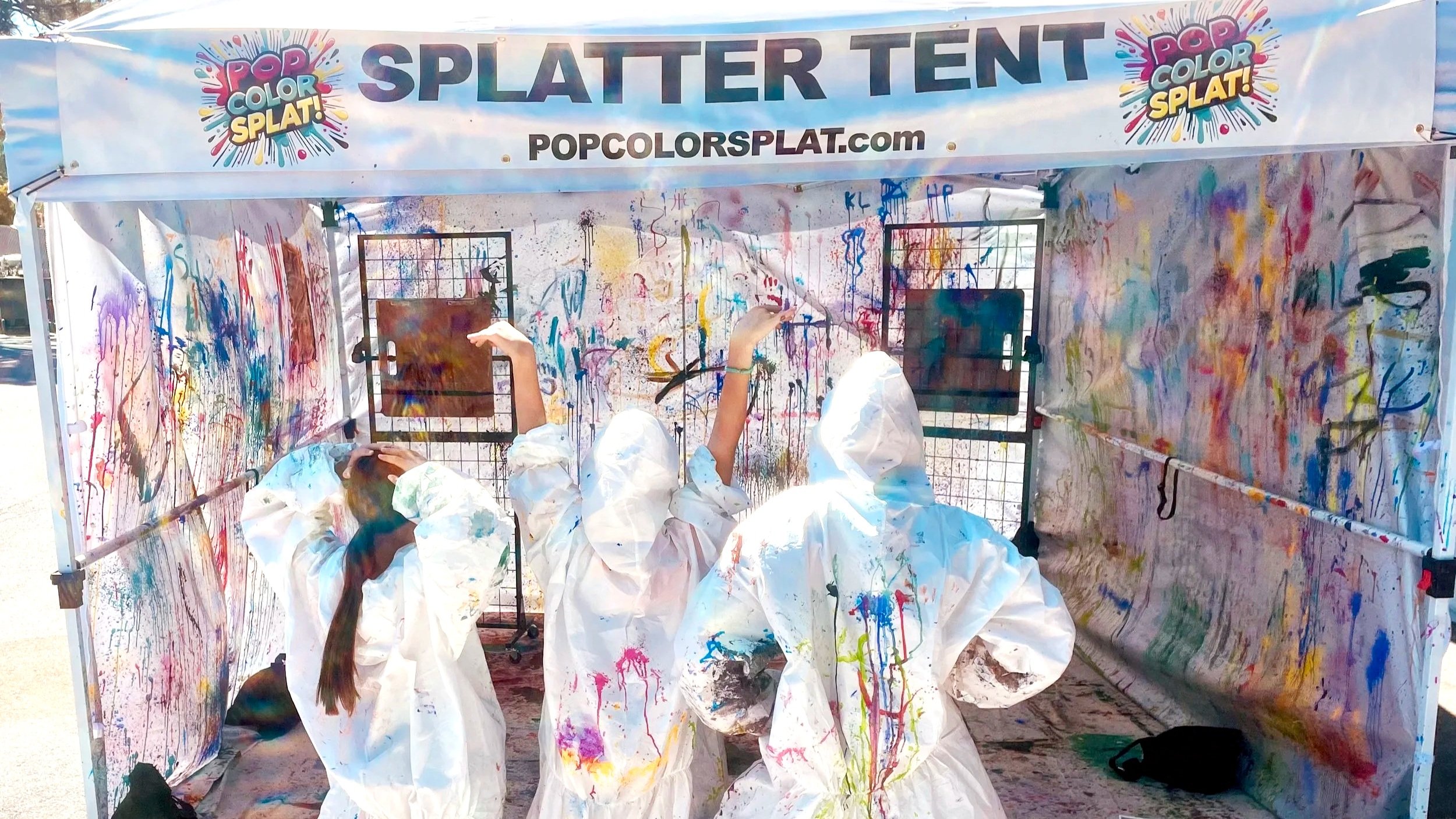 Three teens in protective suits standing in front of a paint splattered Art Tent with a Pop Color Splat Logo and sign that says Splatter Tent.