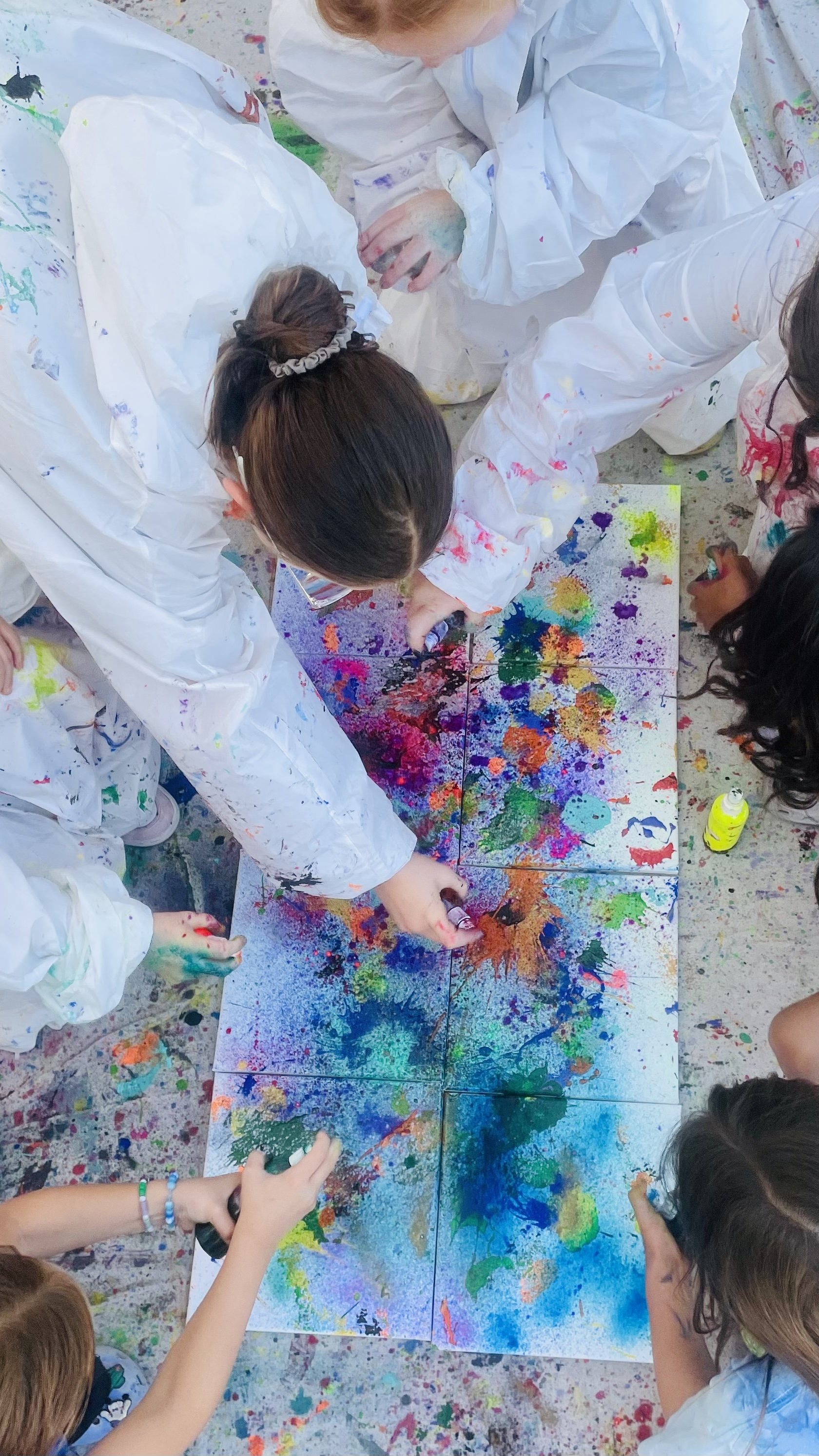 Kids in protective suits gathered around a shared canvas, collaboratively splattering and spraying paint on a large canvas.