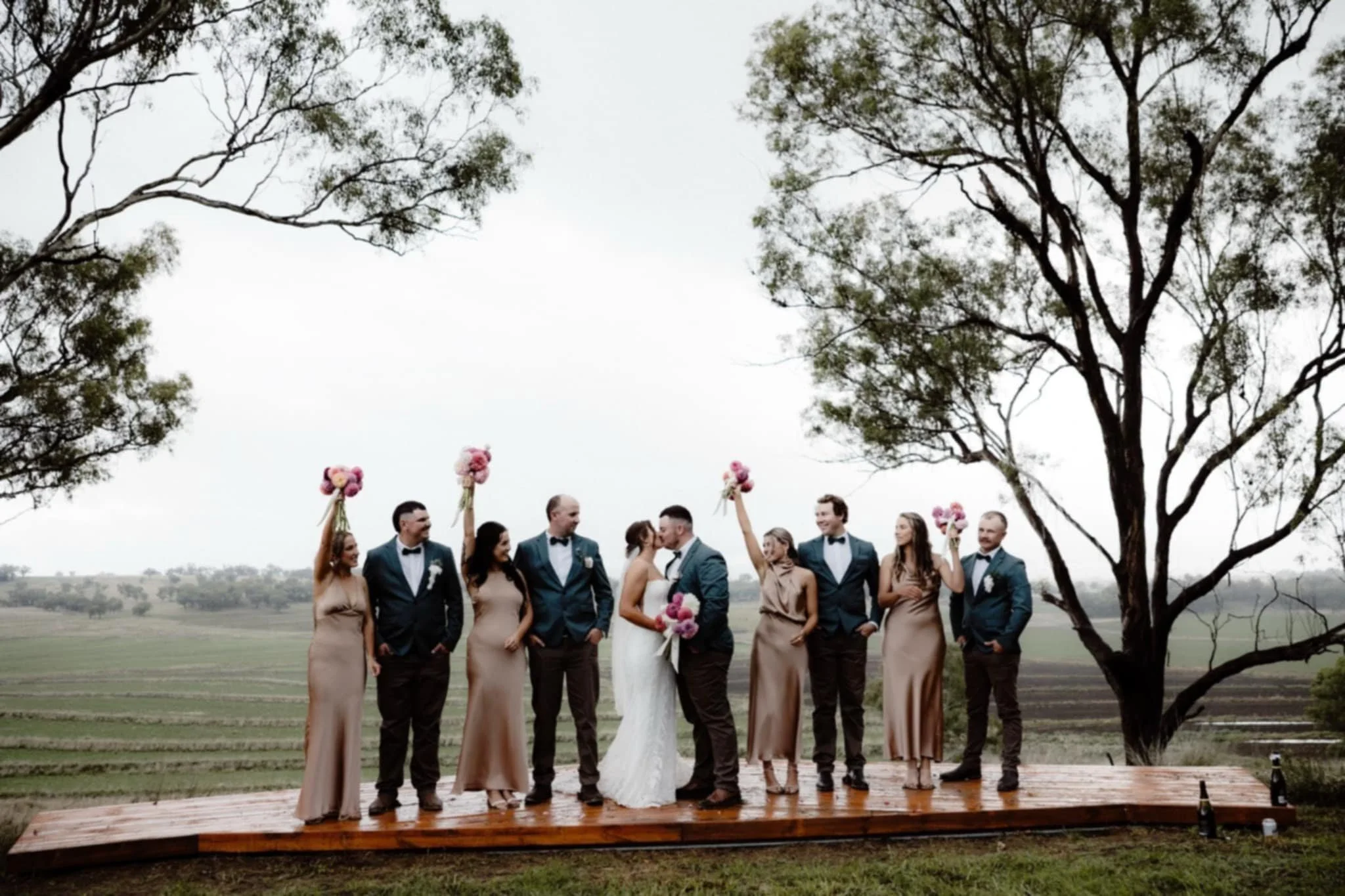 Wedding party on a wooden platform outdoors, with a bride and groom kissing in the center. The group includes bridesmaids in taupe dresses holding pink bouquets and groomsmen in dark suits with bow ties. Trees and fields are in the background.