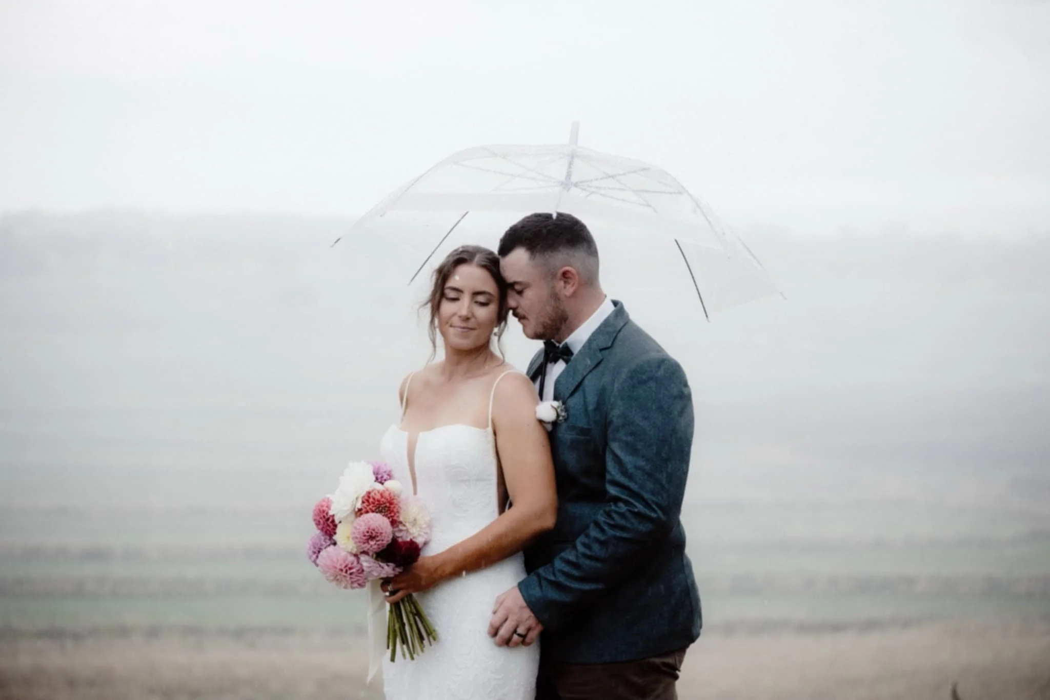 A bride and groom standing close together under an umbrella in the rain, with their foreheads touching and eyes closed, outdoors in a cloudy, rainy environment.