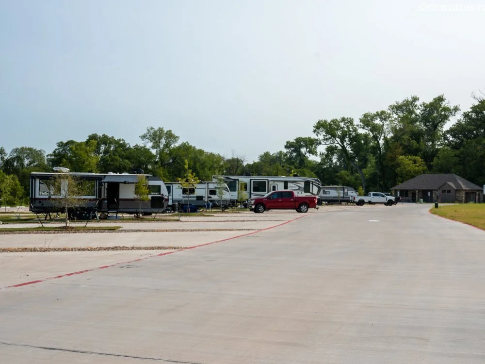 RV parked in a spacious, mostly lot near trees and a small building.