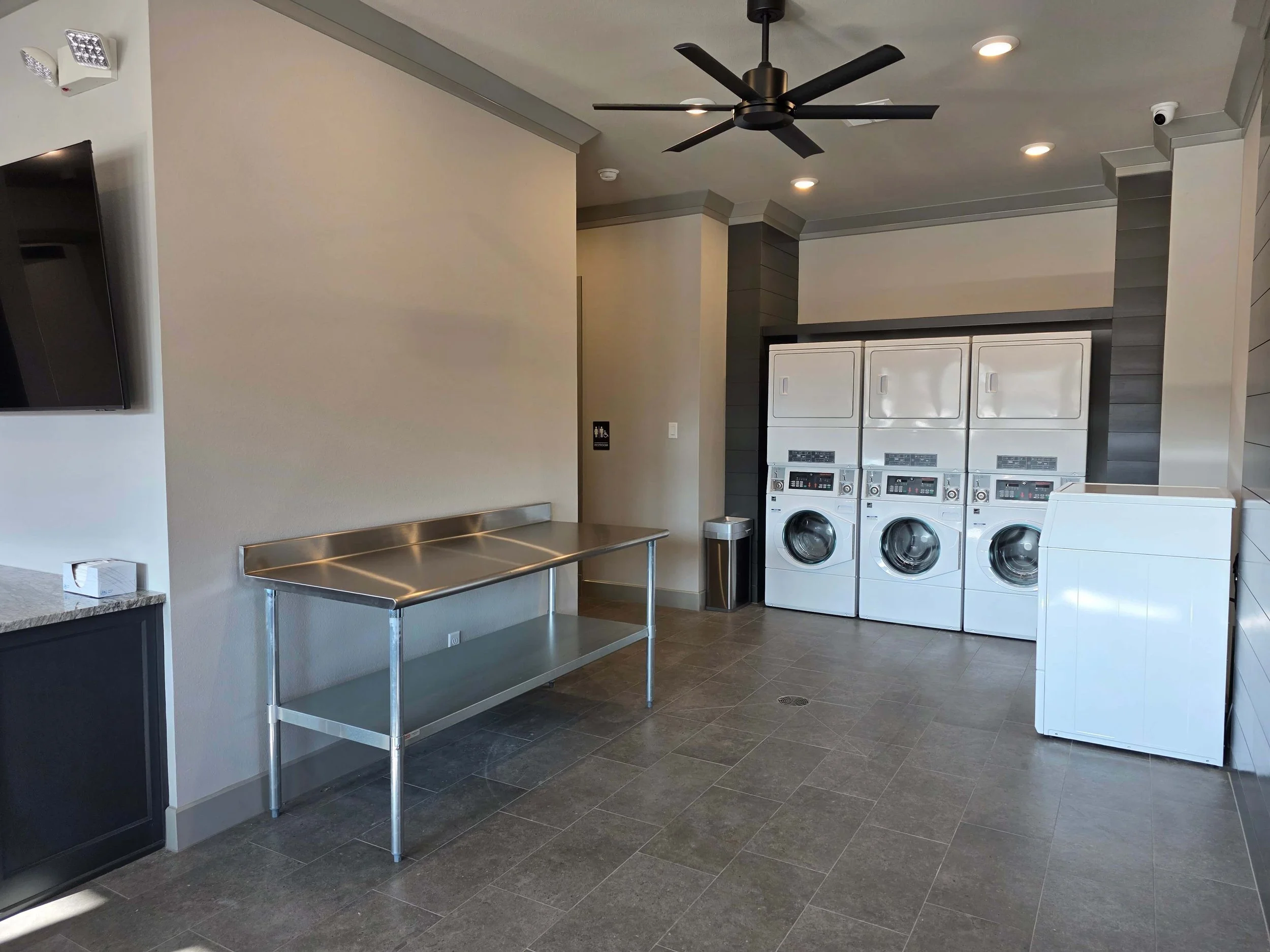 Laundry room with three white industrial washing machines stacked in a row, a black ceiling fan, tiled floor, beige walls, and a stainless-steel table.