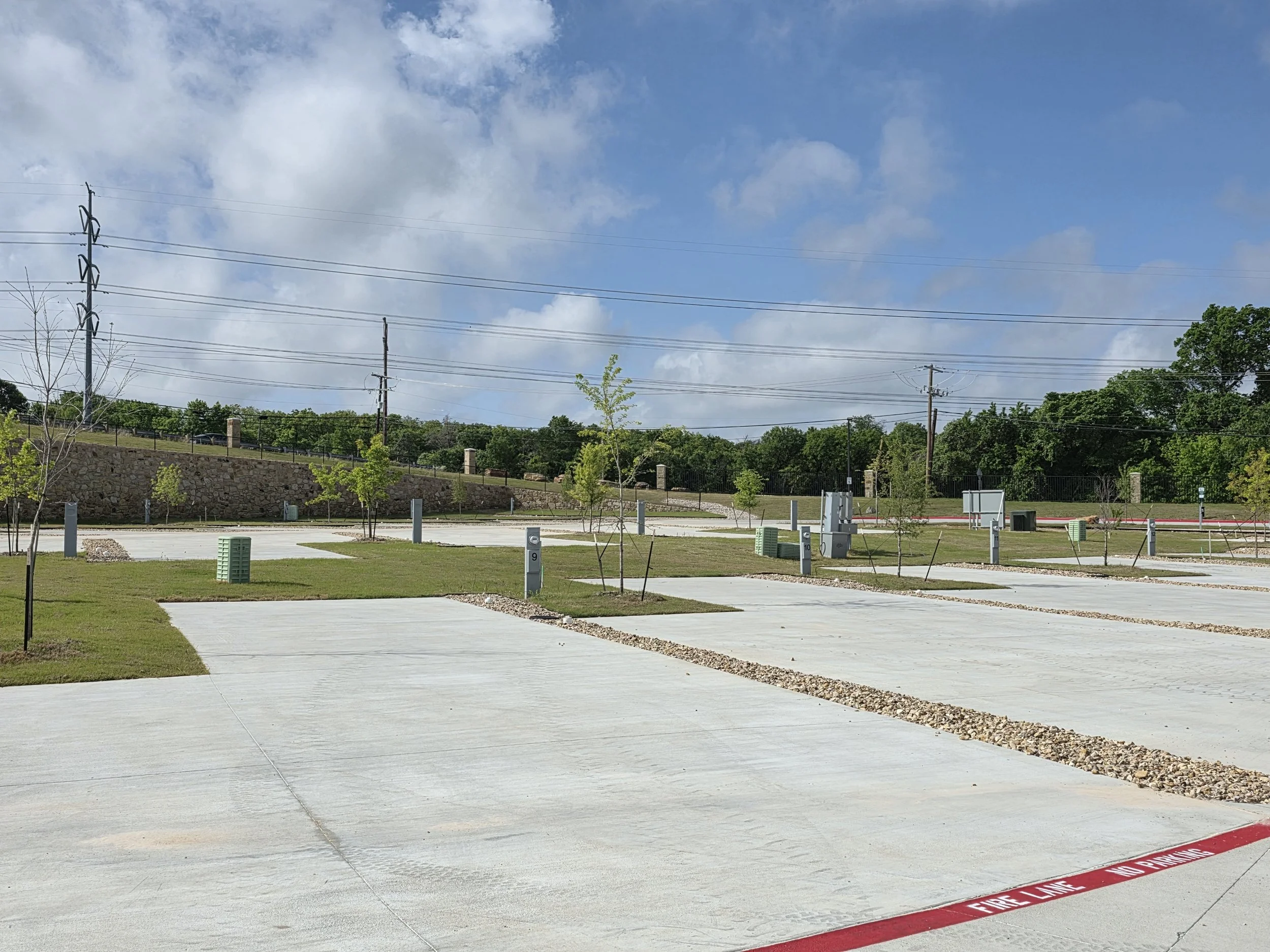 Empty RV lots with concrete pavement, green small trees, electrical poles, and a stone wall with a railing in the background. Blue sky with scattered clouds.