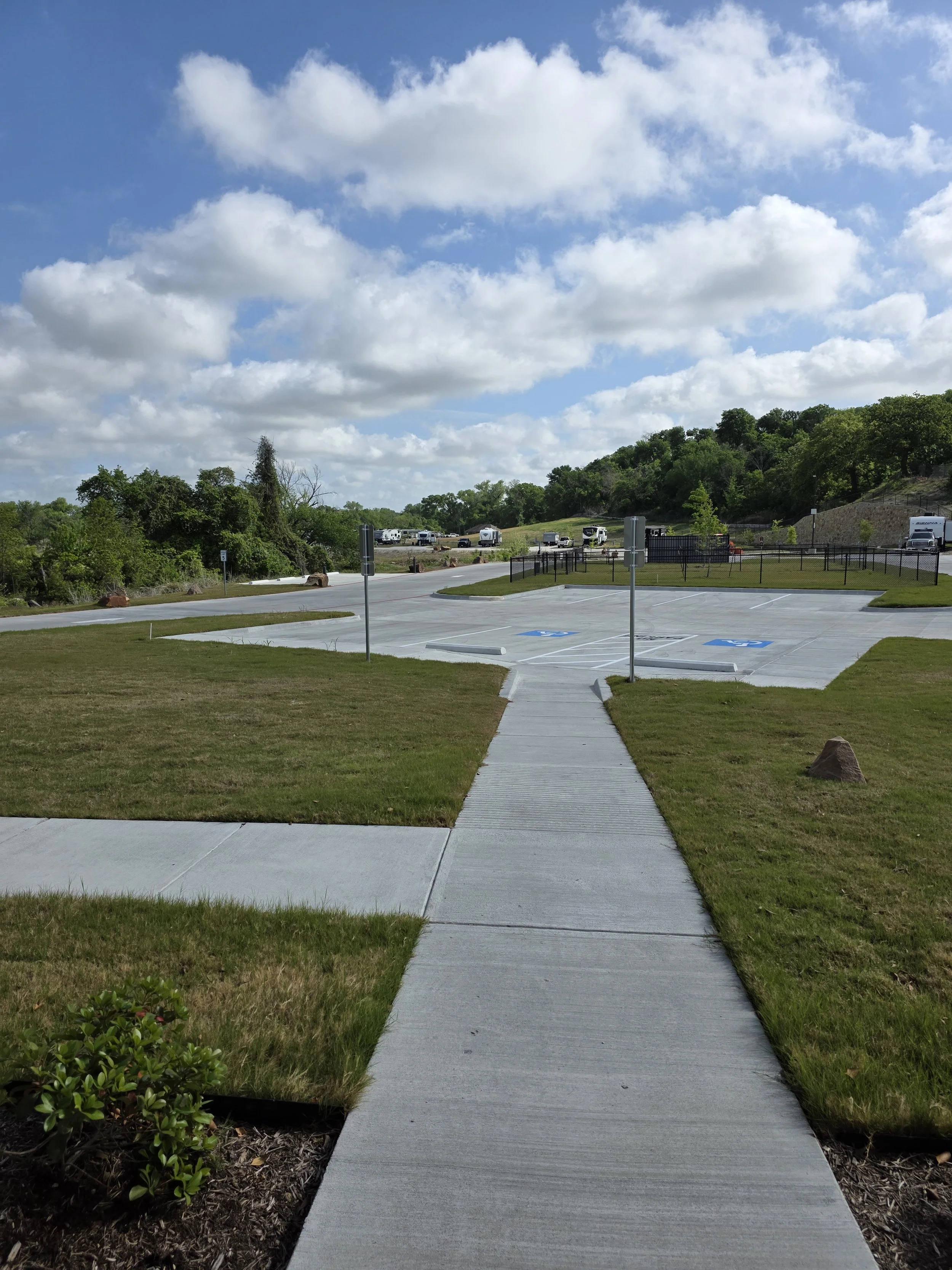 Sidewalk with green grass on all sides, sunny sky, dog park in background.
